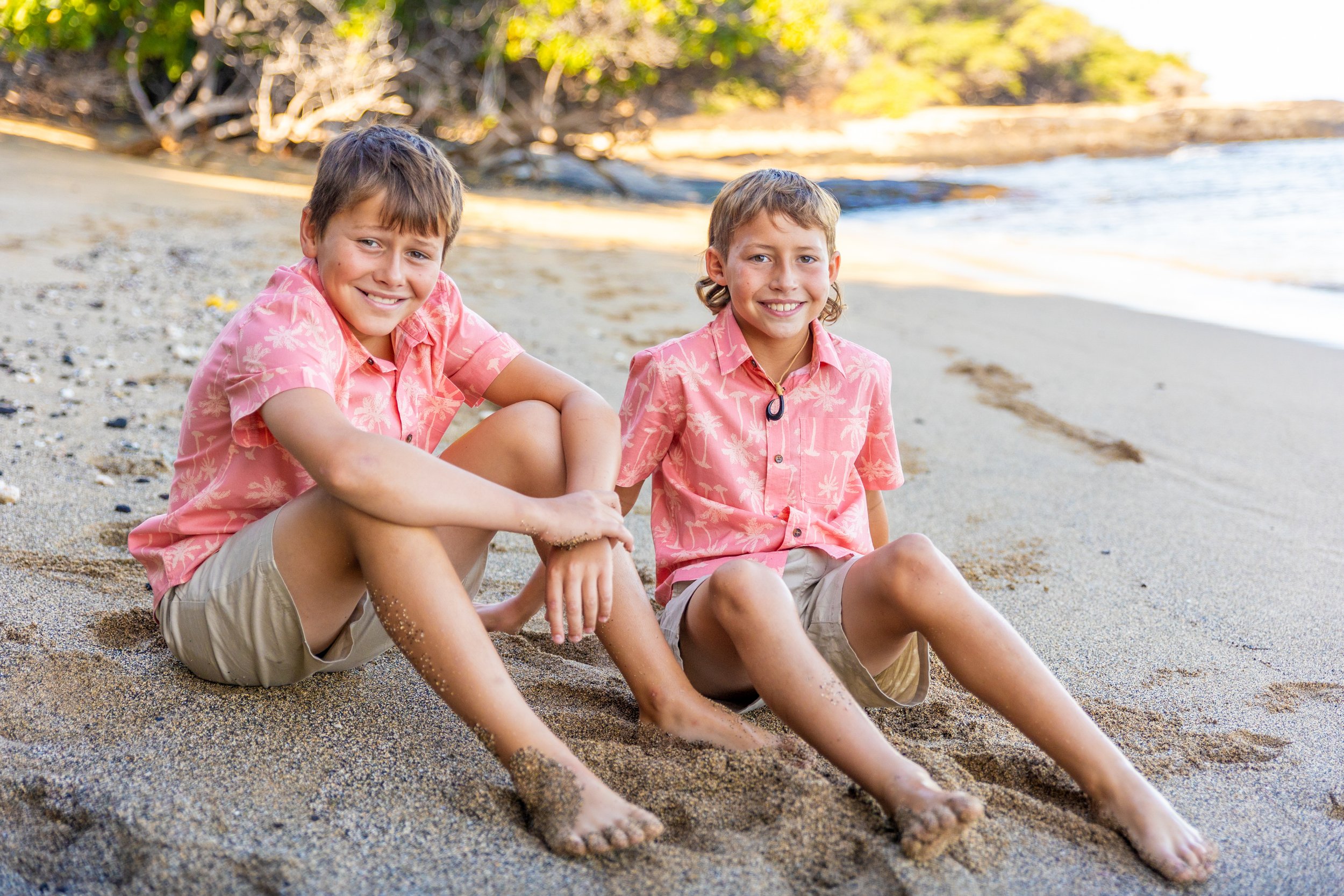 Two young boys sitting on a sandy beach, smiling at the camera, wearing pink Hawaiian shirts and khaki shorts, with shoreline and trees in the background.