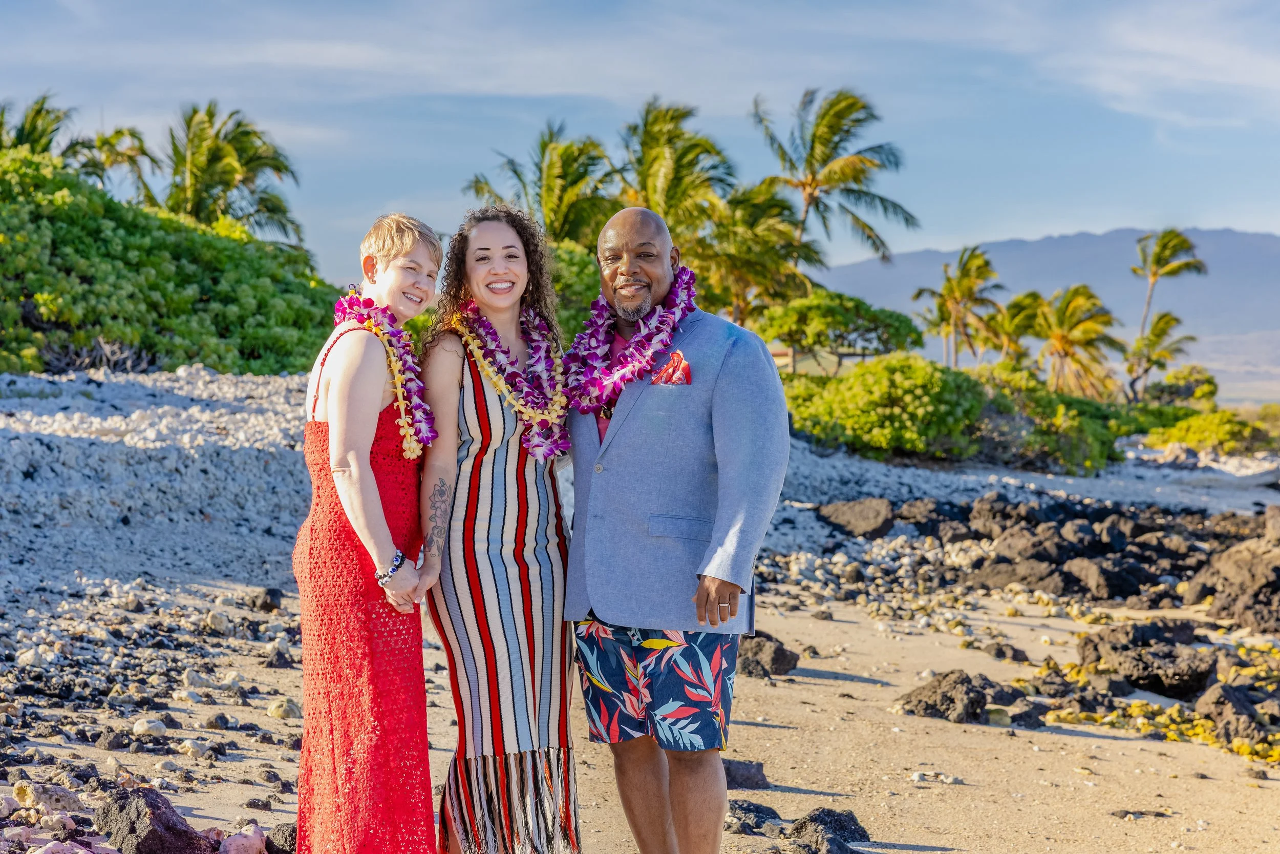 Three people standing together on a rocky beach with palm trees and mountains in the background, wearing leis and smiling.