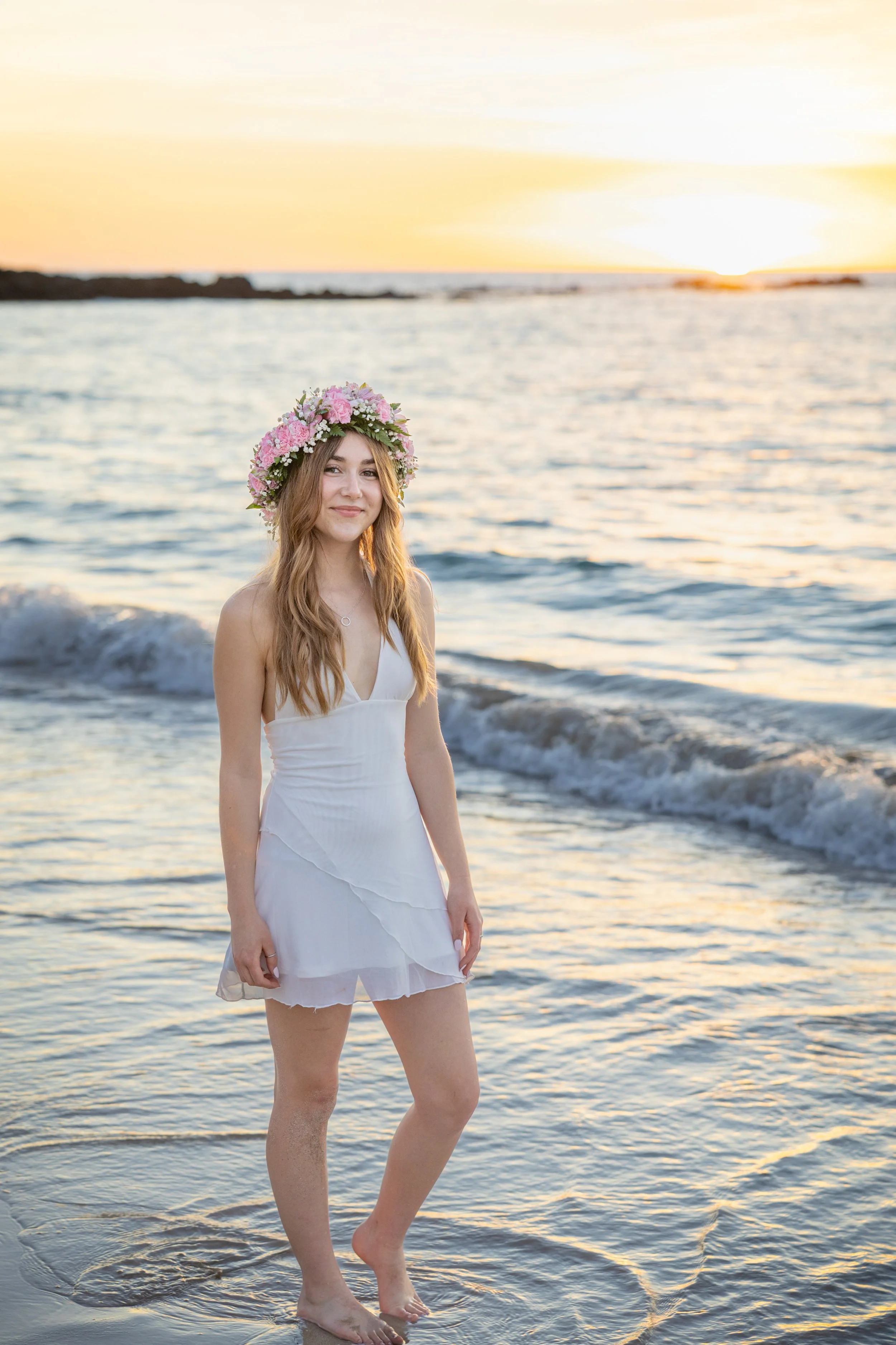 A woman in a white dress and floral crown standing barefoot by the water at sunset.