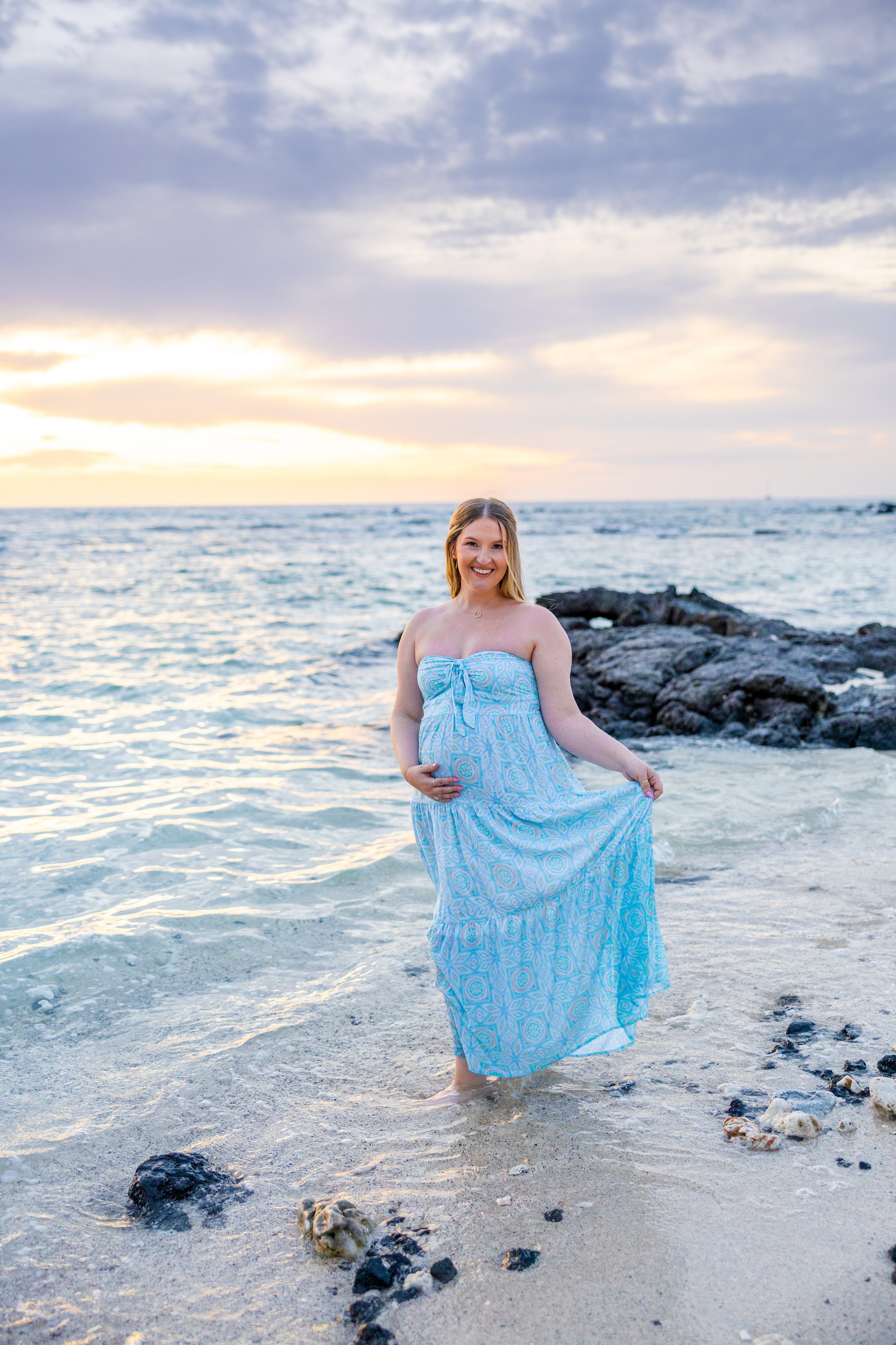 Pregnant woman in a blue dress standing in shallow ocean water at sunset.