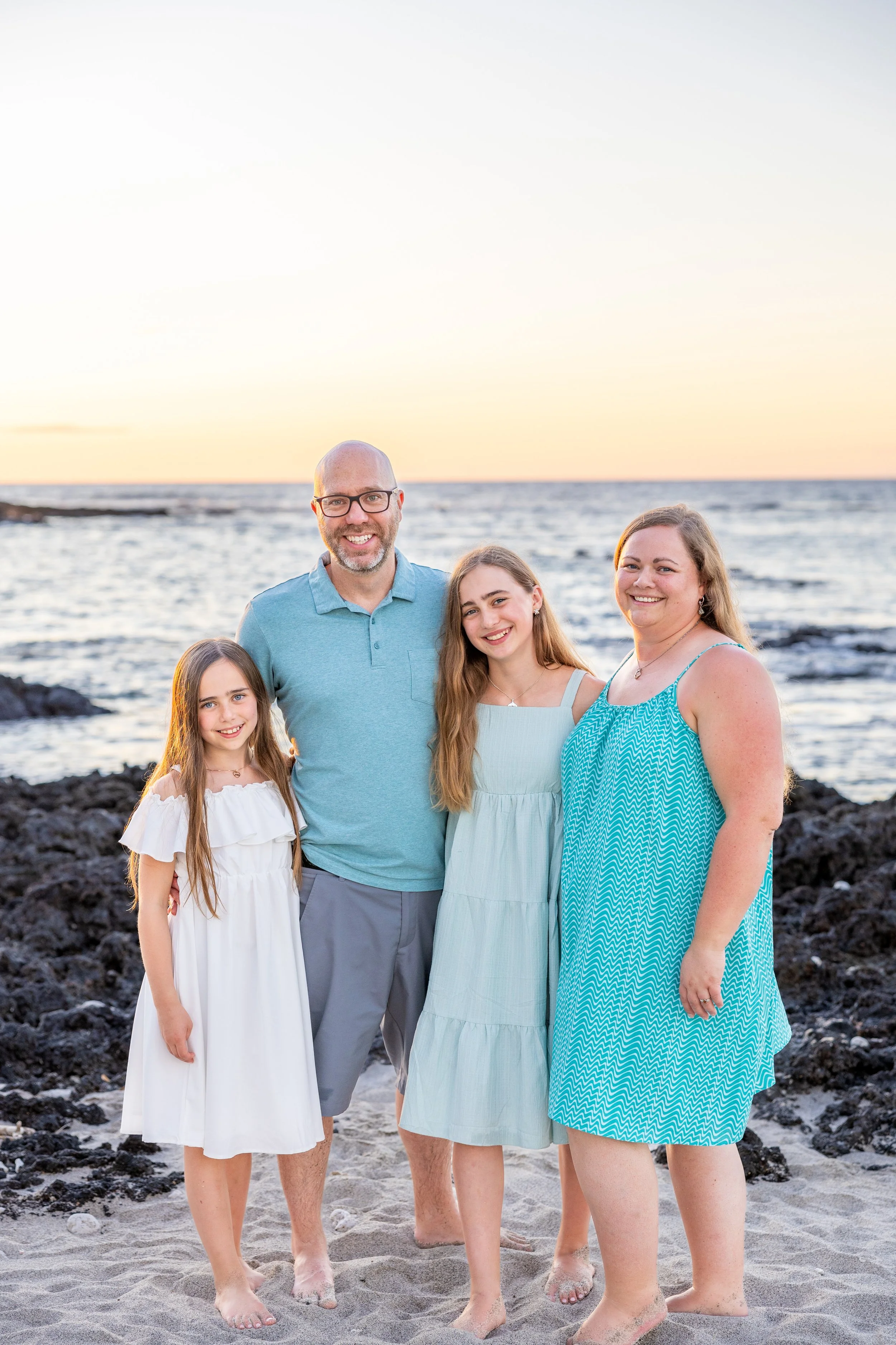A family of four standing on a rocky beach at sunset, smiling at the camera. The family consists of a man, a woman, and two girls, all dressed in light-colored summer clothing.