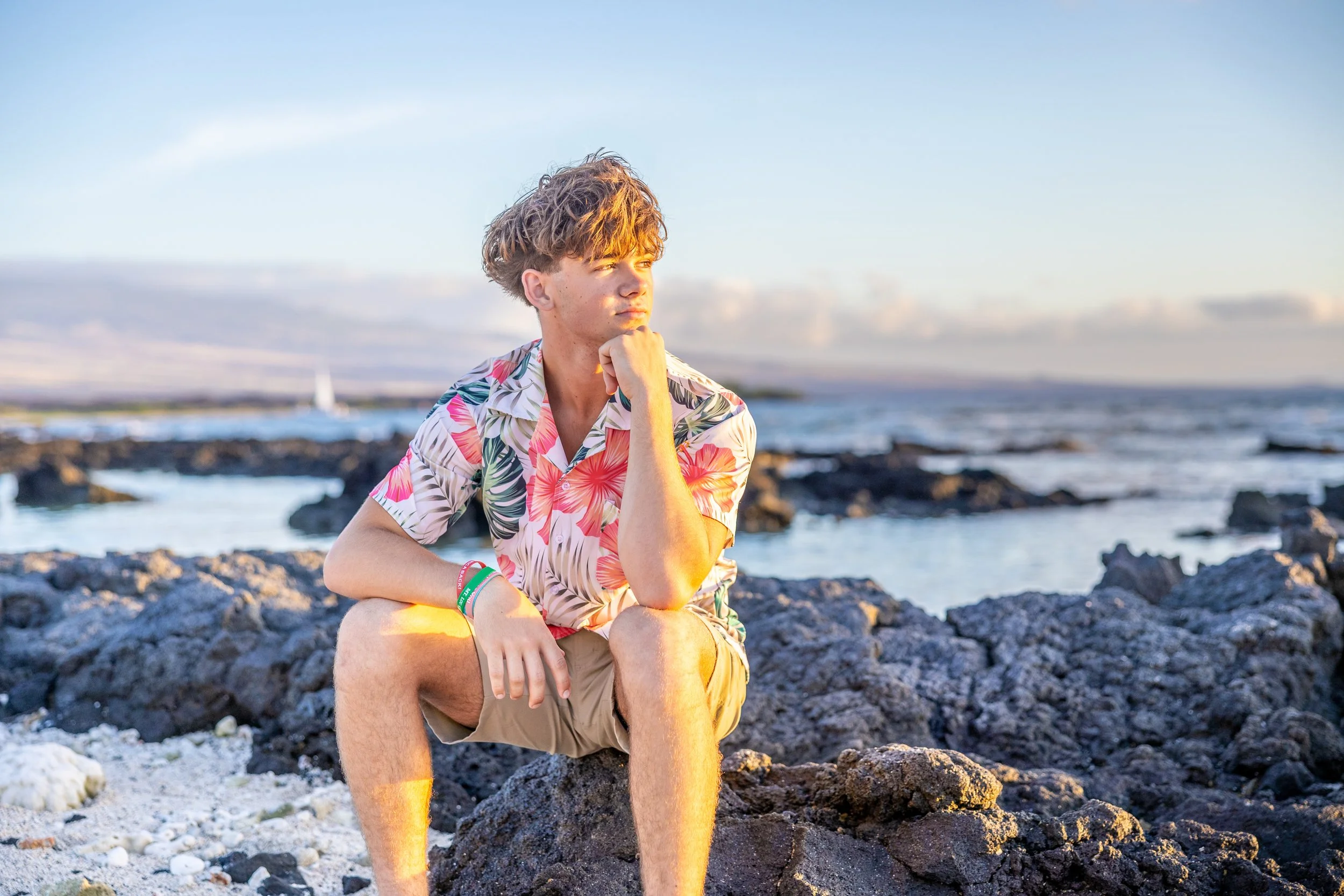 A young man sitting on rocks by the ocean, wearing a colorful tropical shirt and beige shorts, looking thoughtfully into the distance during sunset.