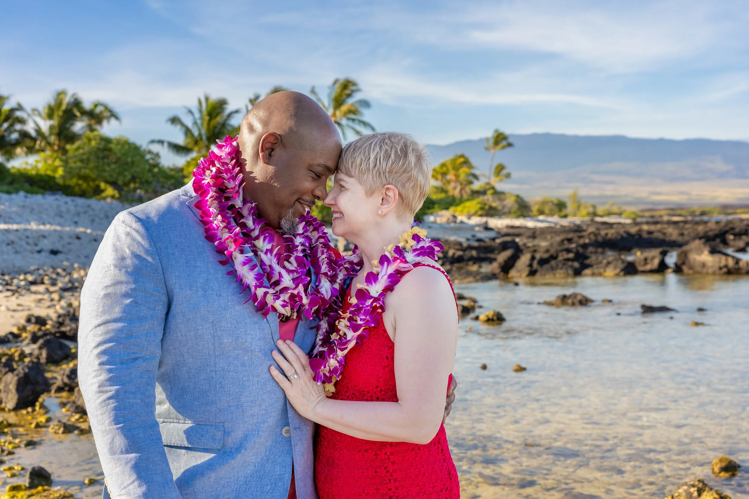 A romantic couple stands close together on a beach, touching foreheads and smiling. The man is wearing a light gray suit, and the woman is in a red dress. Both are wearing purple lei necklaces. The background features palm trees, rocks, water, and a 