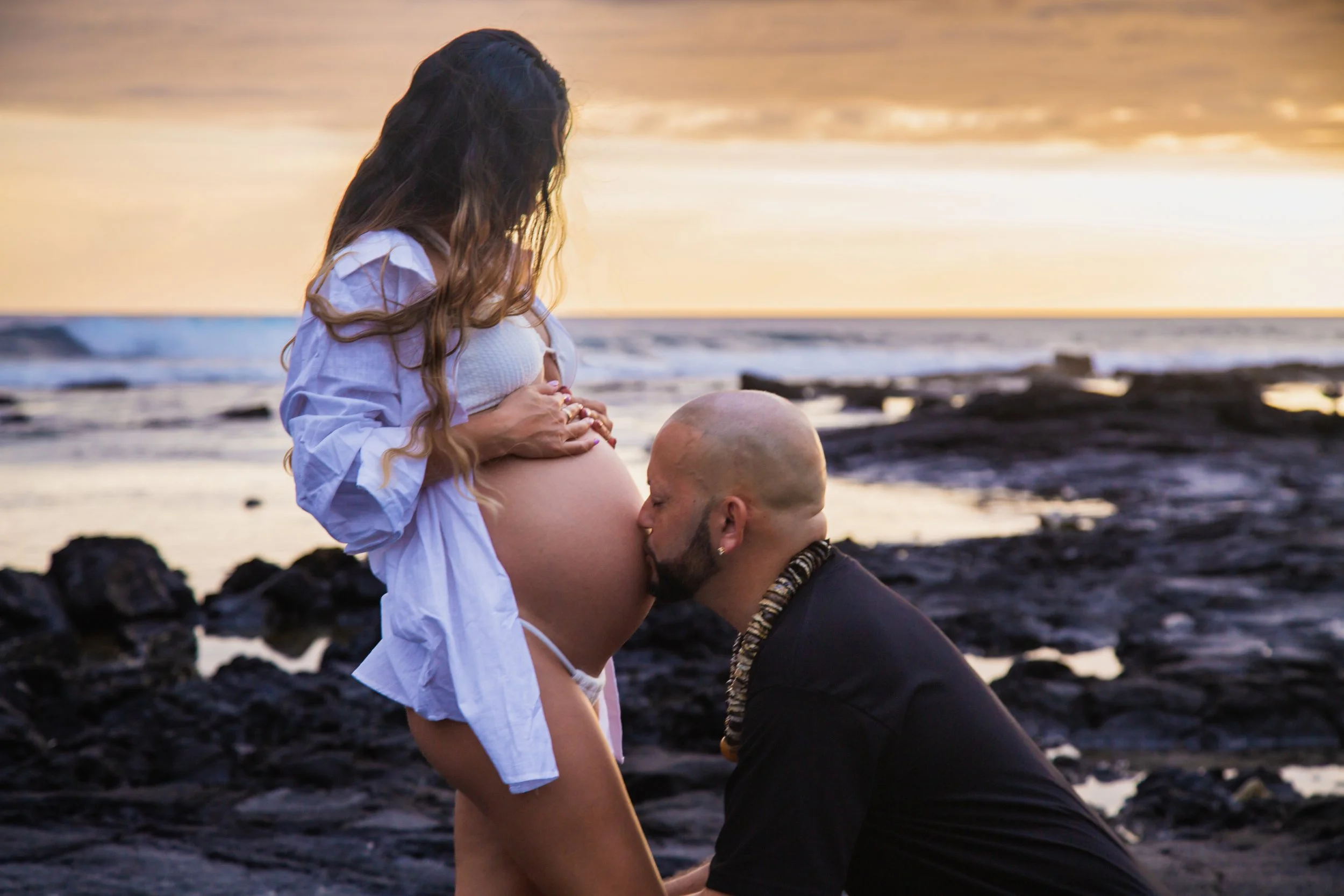 A pregnant woman standing on a rocky beach at sunset, holding her belly. A man kneeling in front of her is kissing her belly.