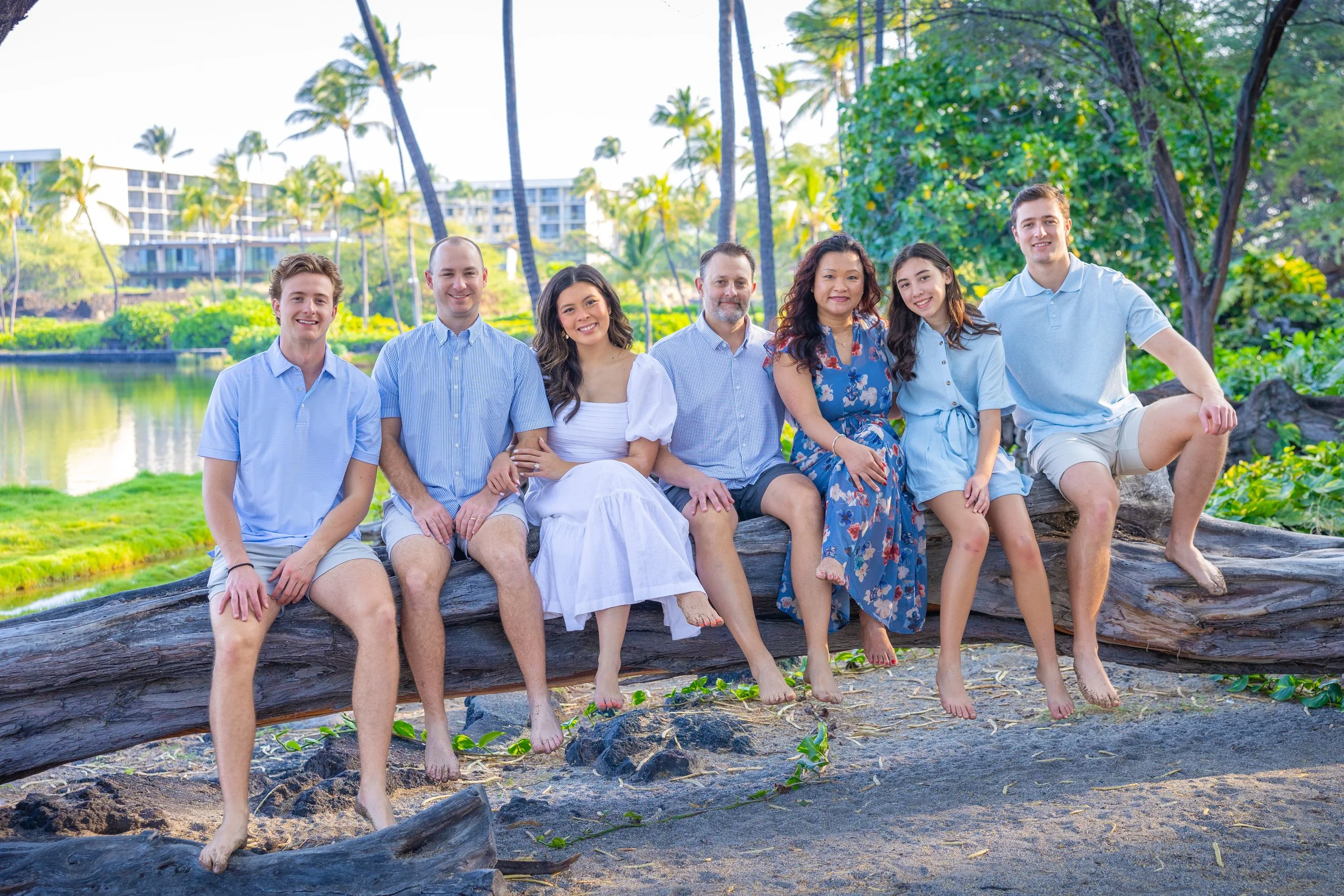 Group of seven people sitting on a large log on a beach, with palm trees and buildings in the background, smiling and enjoying a sunny day.