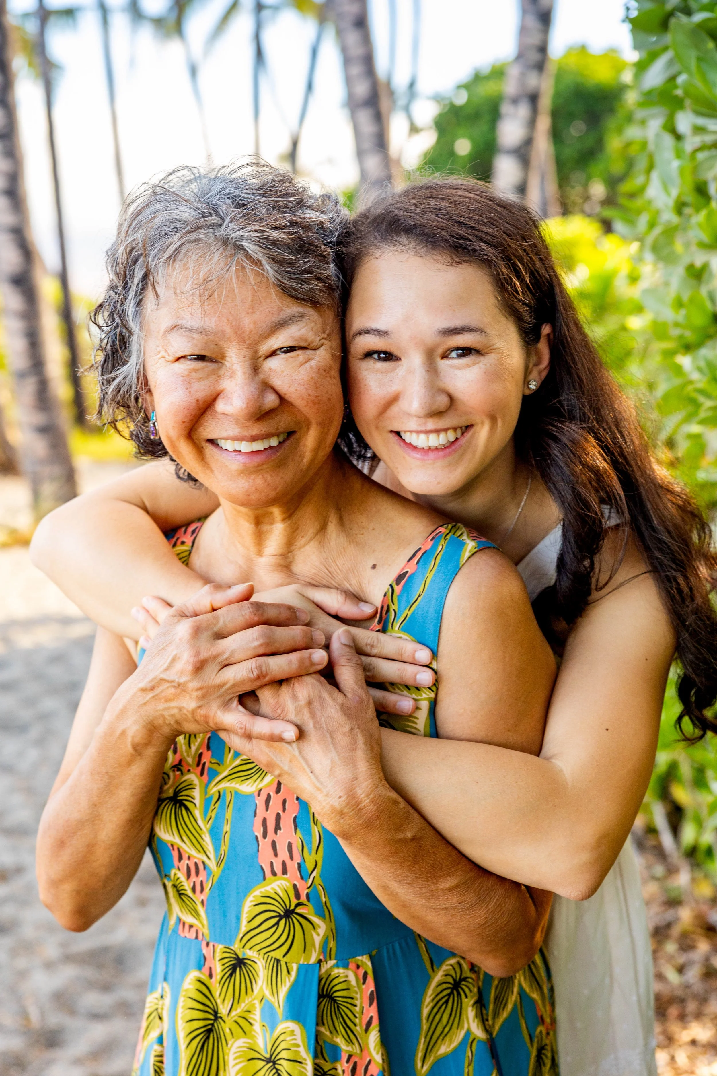 Two women, an older woman and a younger woman, smiling and hugging outdoors with tropical trees and greenery in the background.