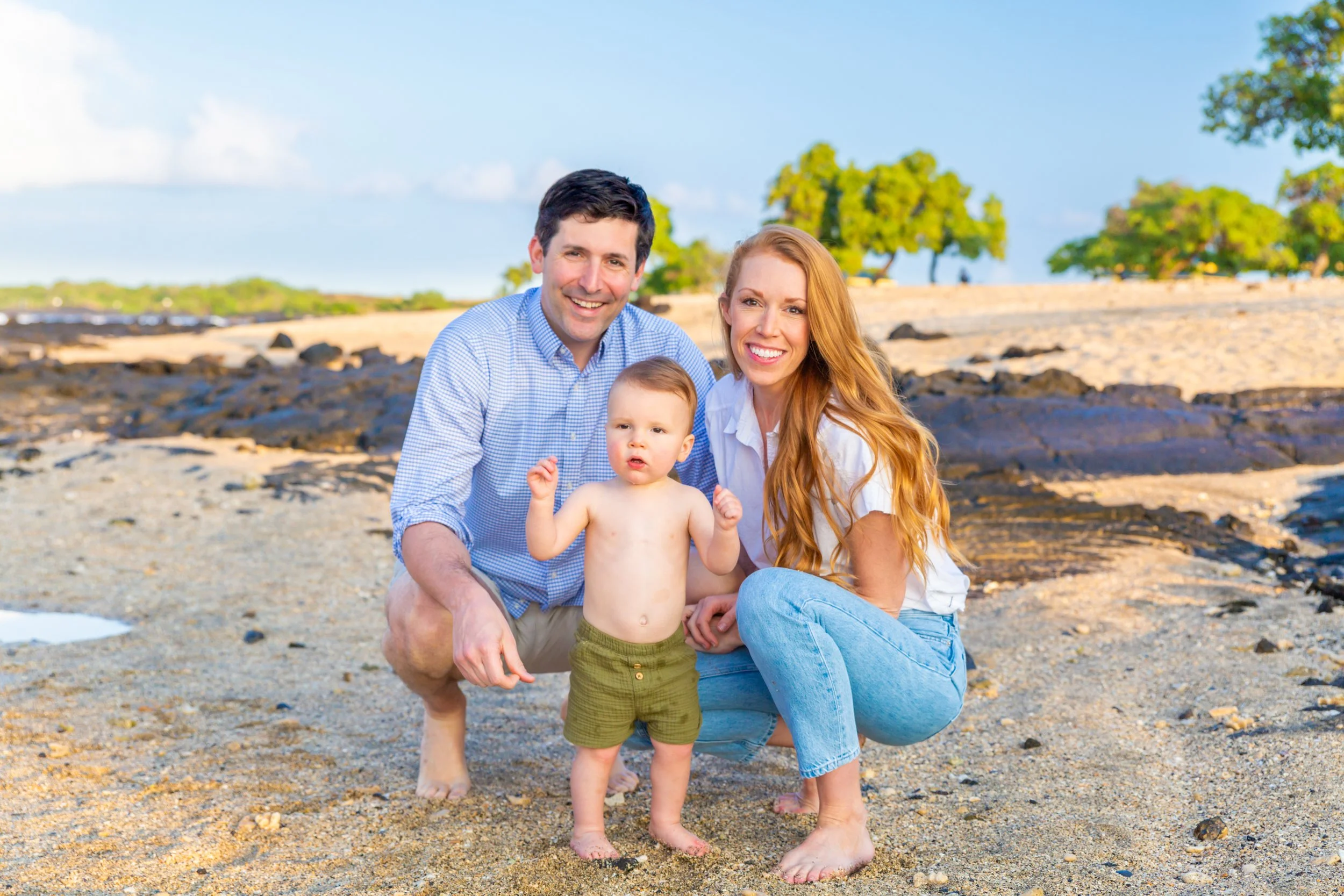 A happy family of three, consisting of a man, a woman, and a toddler, on a sandy beach with rocks and green trees in the background. They are all smiling, with the toddler in the center and the parents kneeling beside the child.