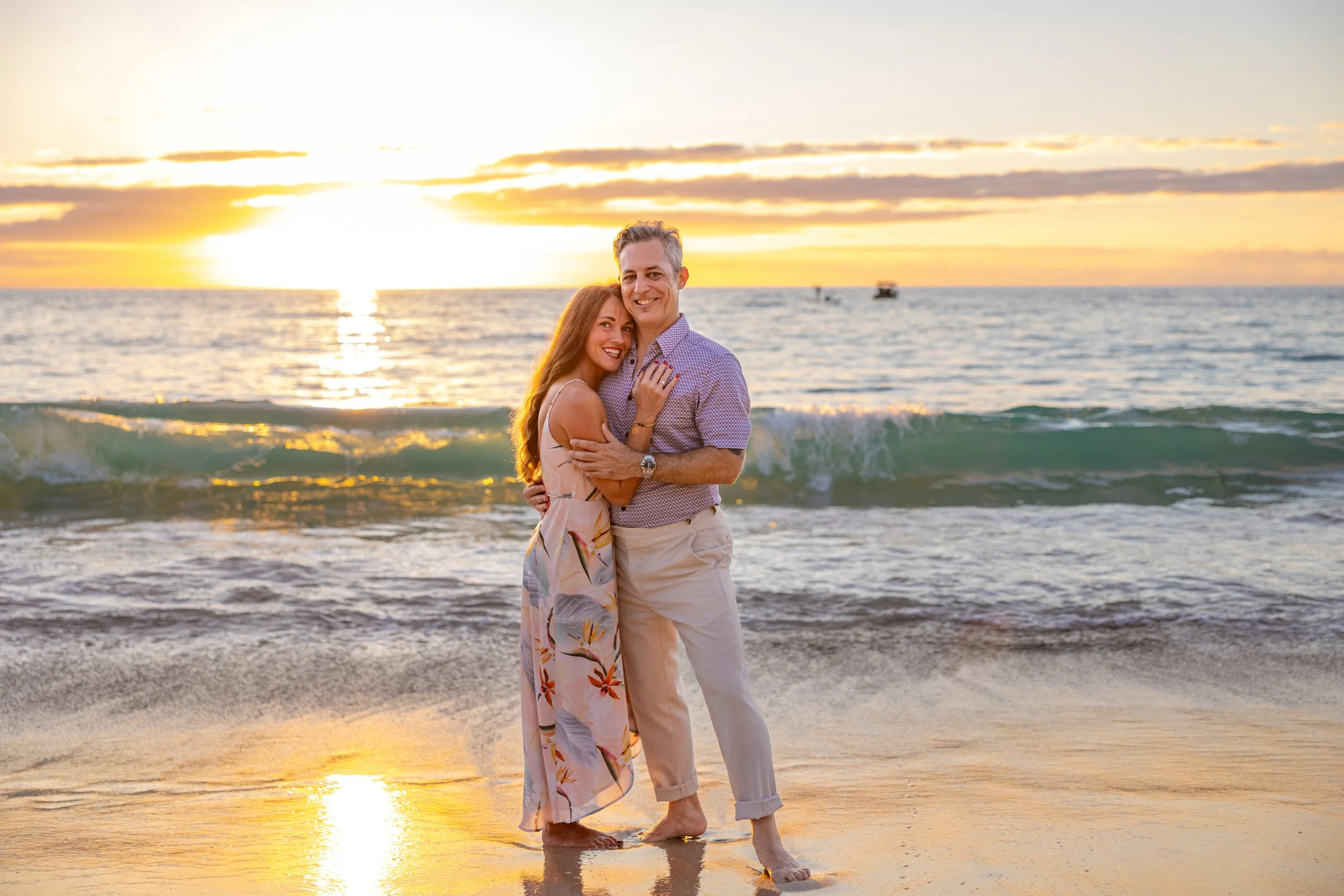 A happy couple stands on the beach with the ocean and sunset in the background. The woman has long red hair and wears a floral dress, while the man has short gray hair and wears a purple patterned shirt with beige pants.