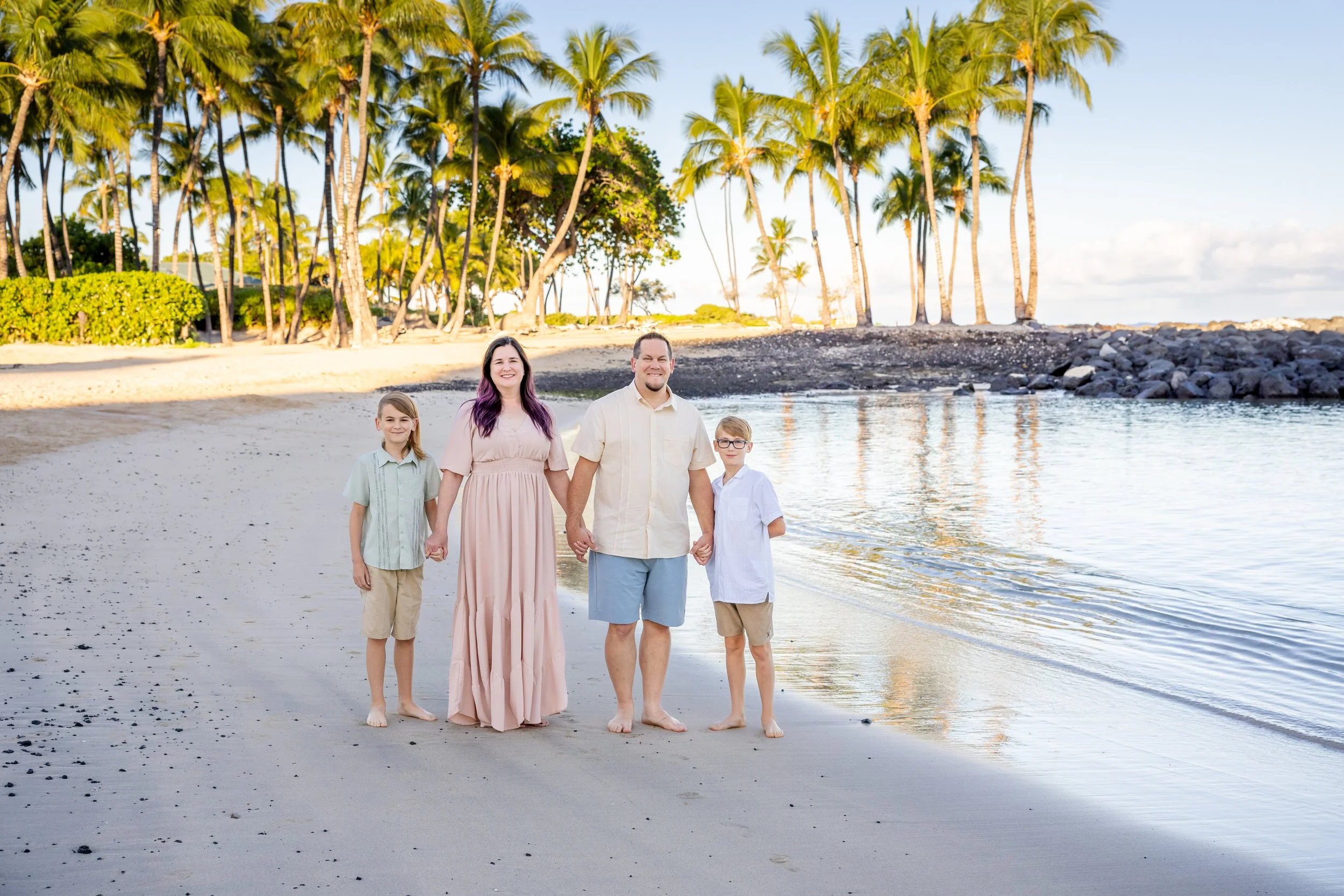 A family of four holding hands and walking on a beach with palm trees in the background.