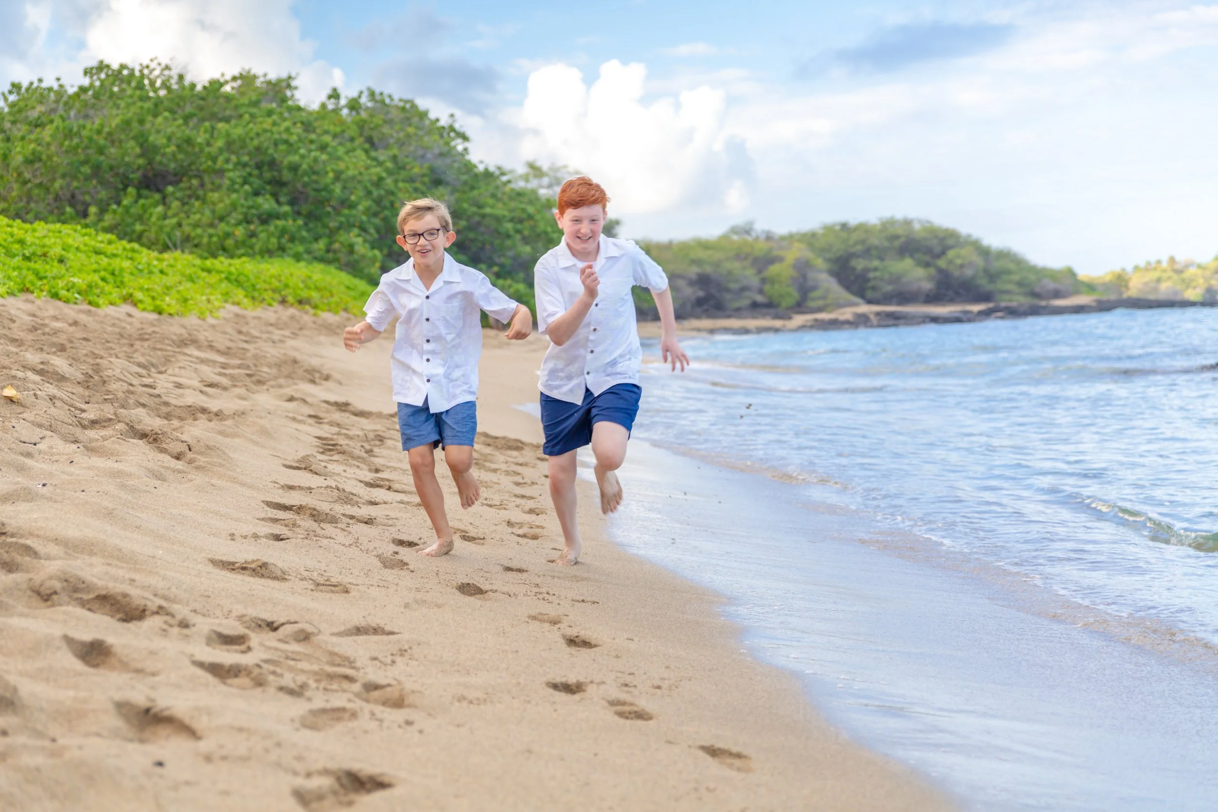 Two young boys running barefoot on a sandy beach near the water with lush green foliage and a cloudy sky in the background.