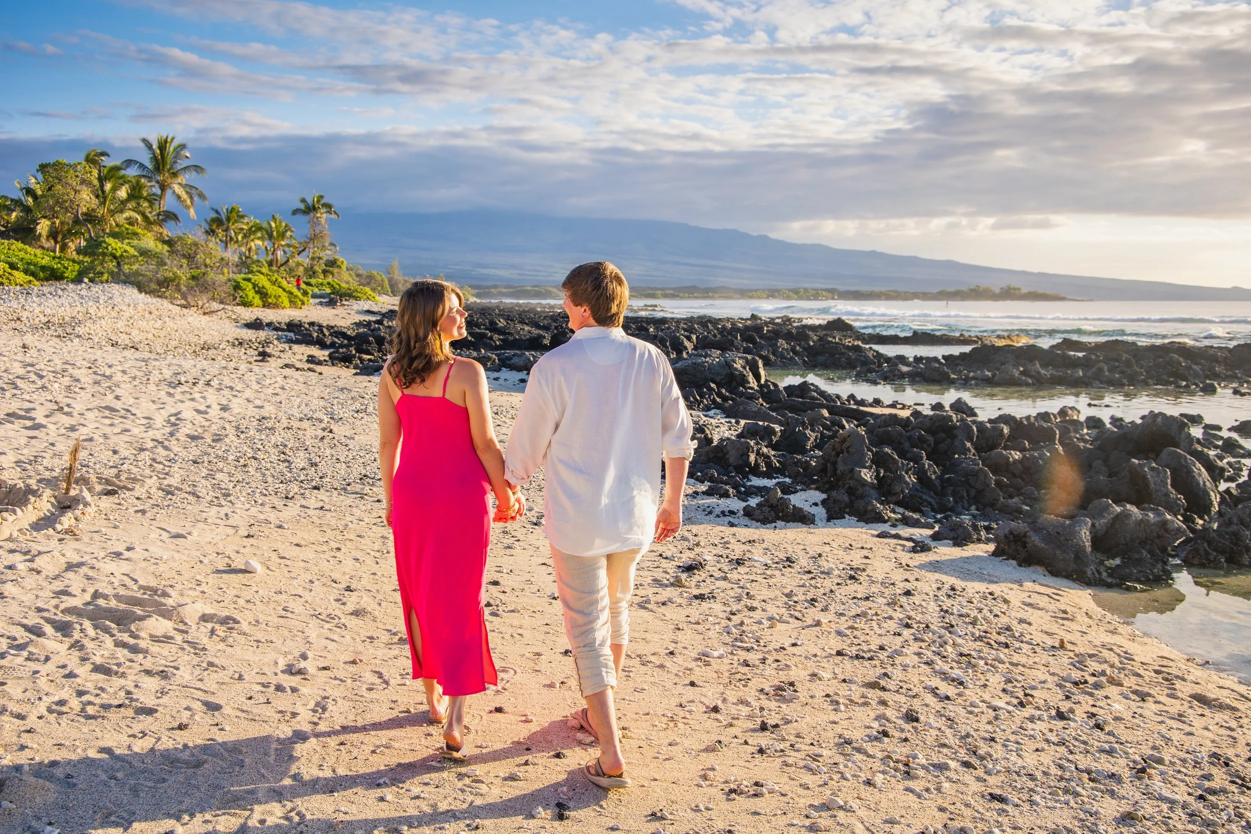A man and woman holding hands walk along a sandy beach with rocks, palm trees, and ocean in the background during sunset.