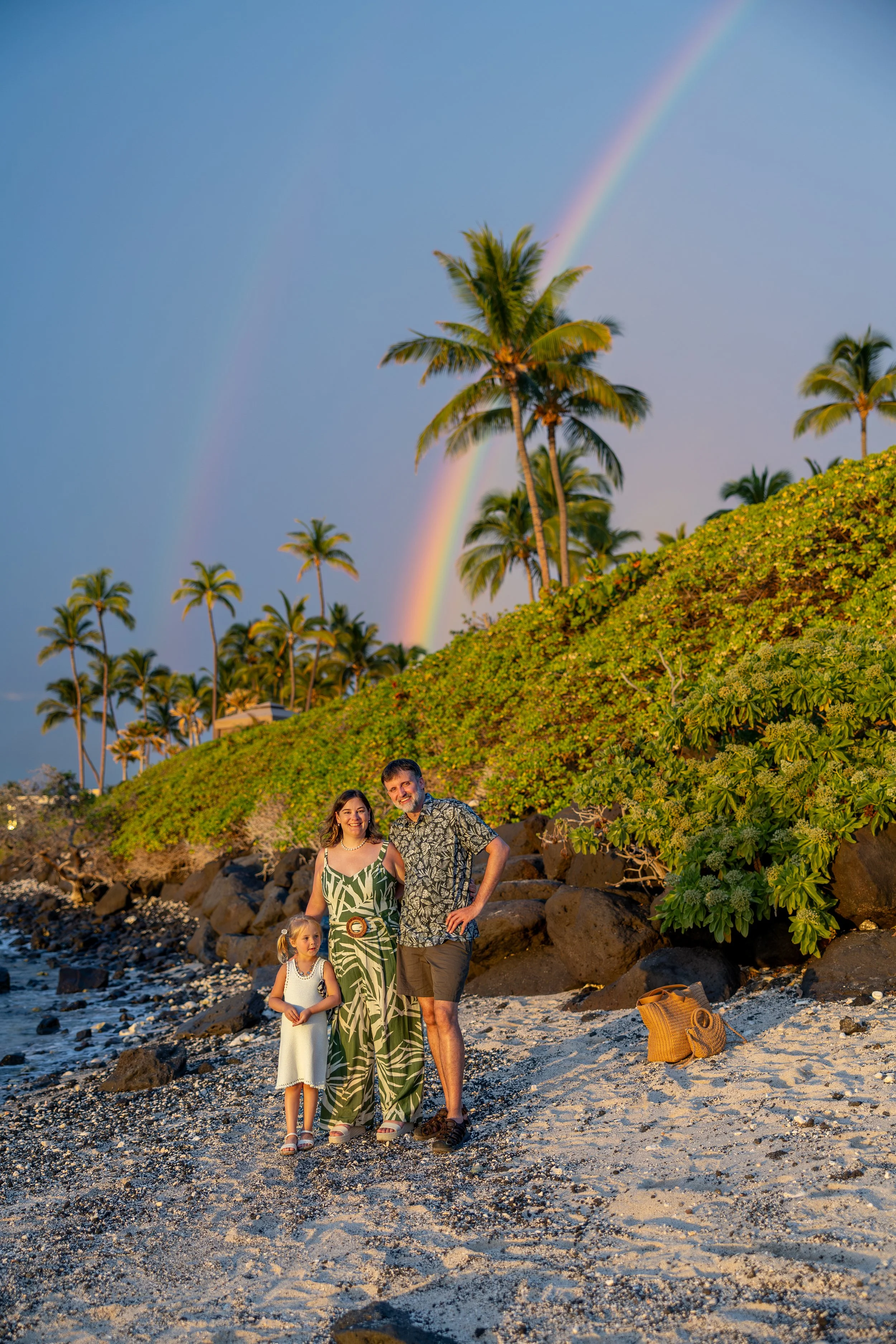 Family of three, a woman, a man, and a young girl, standing on a beach with palm trees and a rainbow in the background during sunset.
