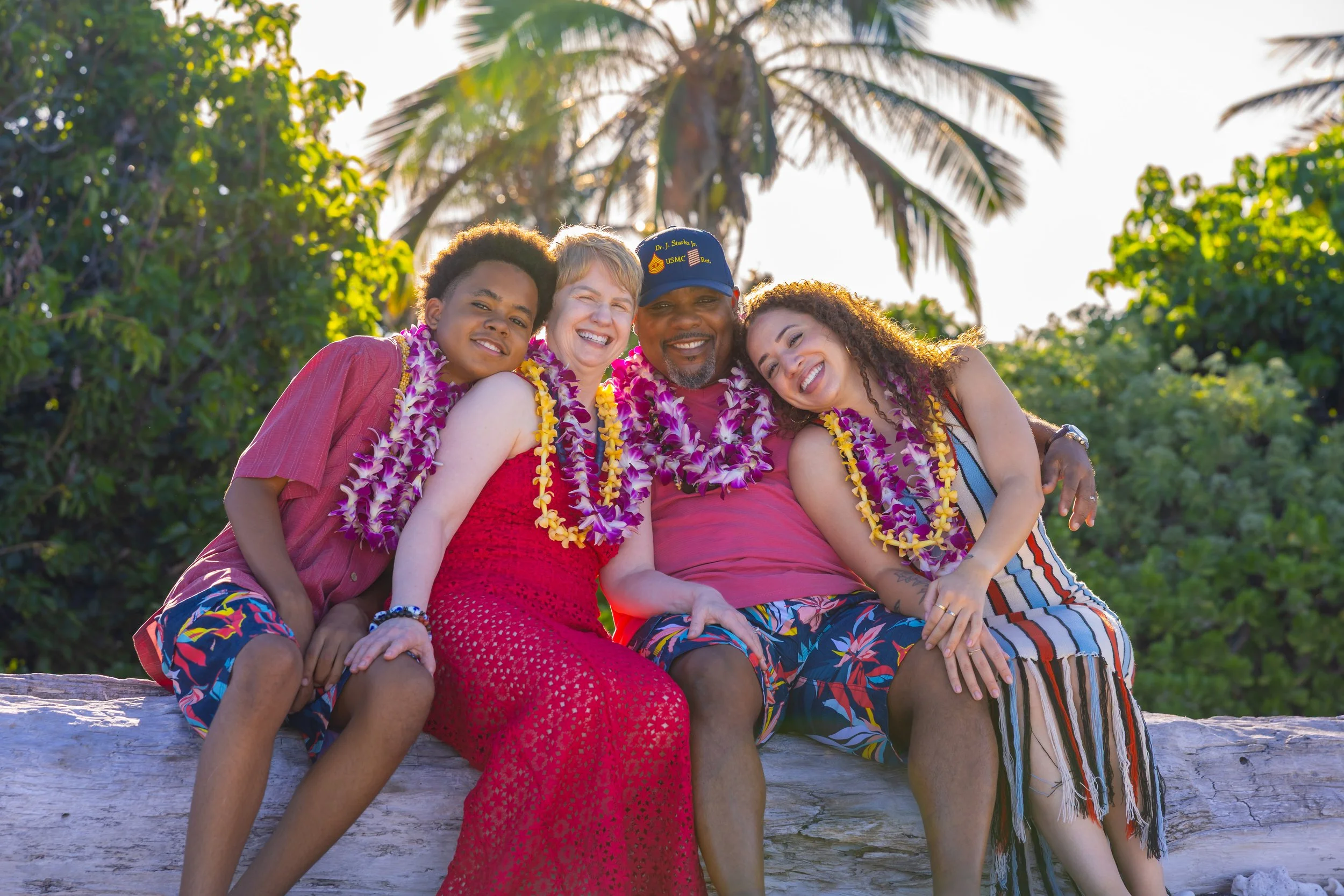 A happy family of four sitting on a log, wearing leis, with palm trees and greenery in the background, celebrating a vacation or special occasion in a tropical location.