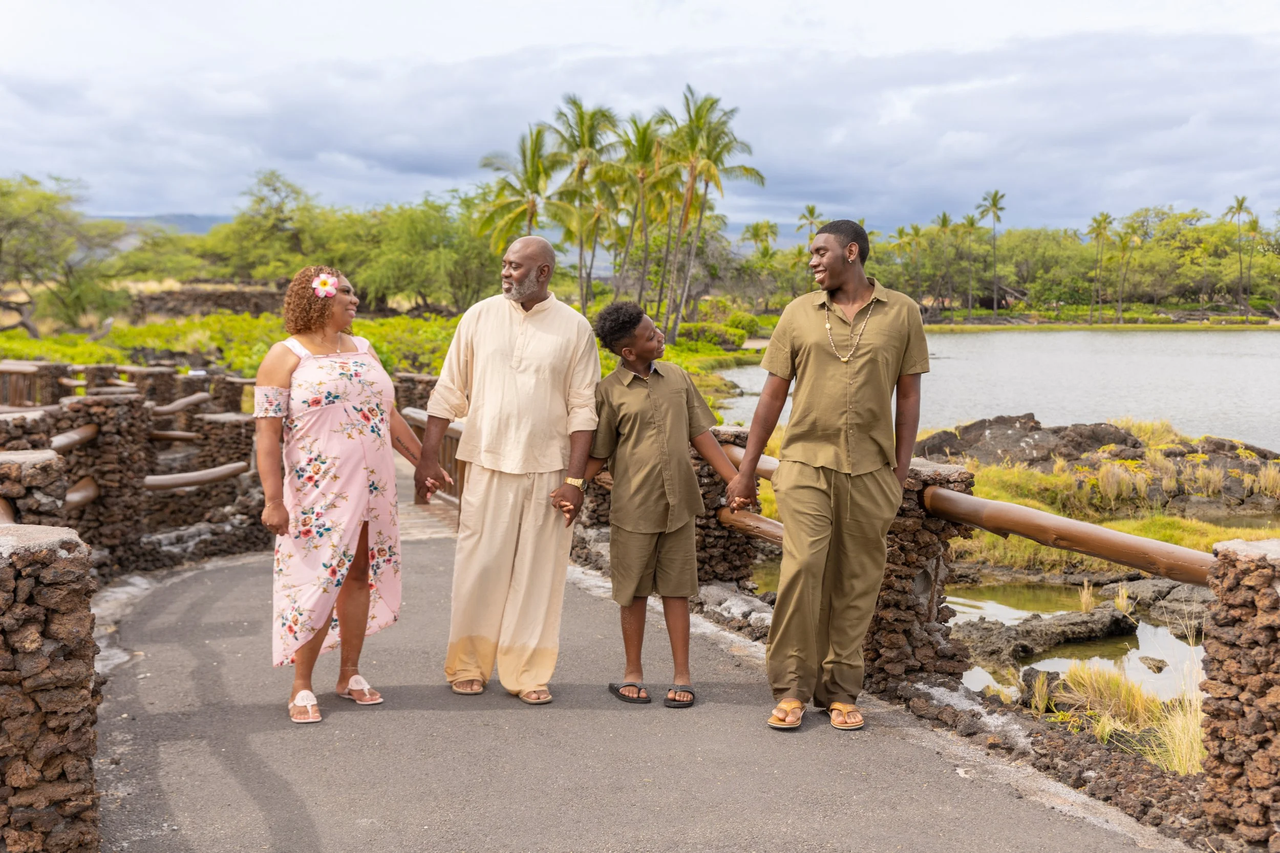 A family of five holding hands while walking on a stone pathway near a lake with palm trees and lush greenery in the background.