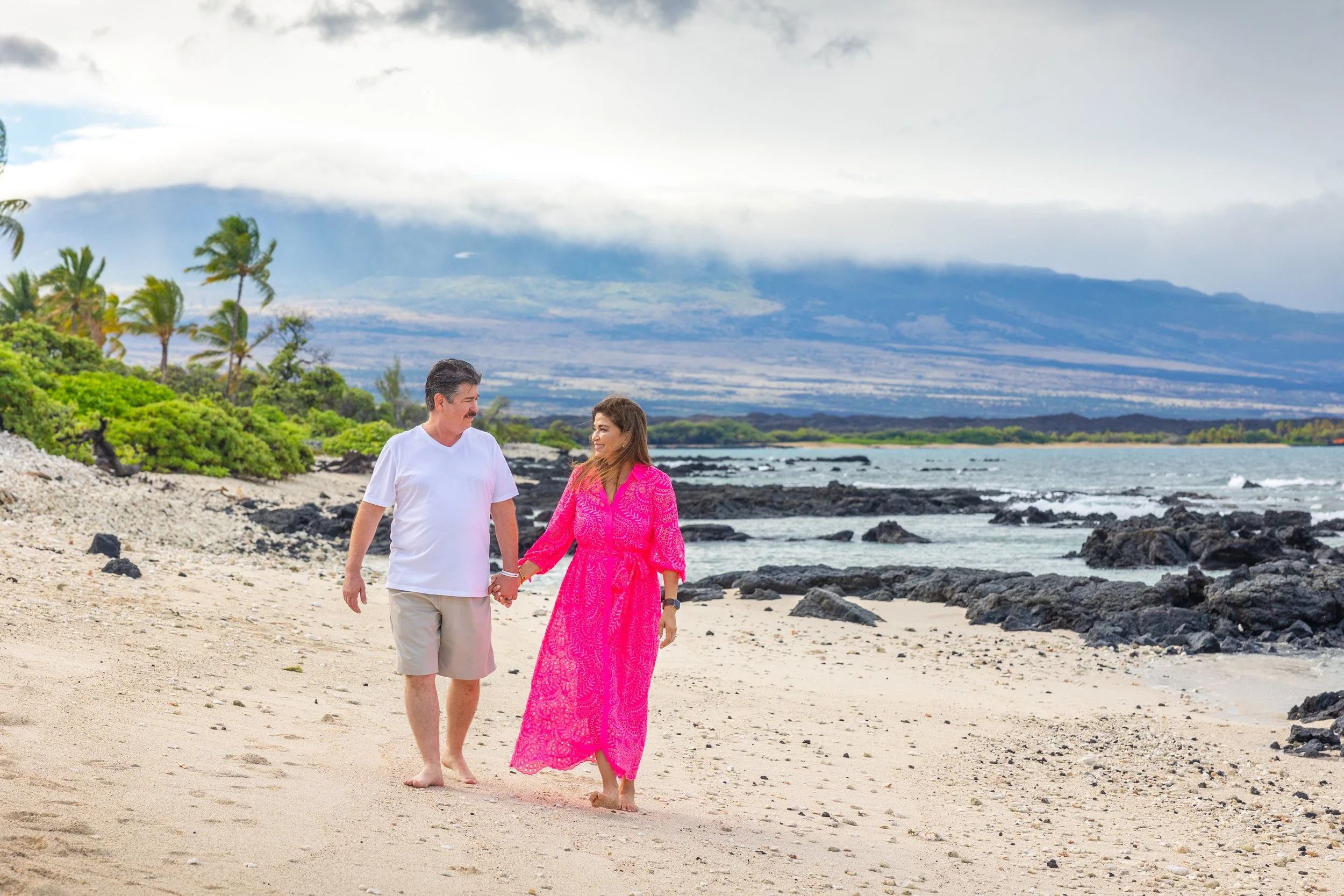 A couple walking hand in hand along a sandy beach with black rocks, lush green foliage, and mountains in the background under a partly cloudy sky.