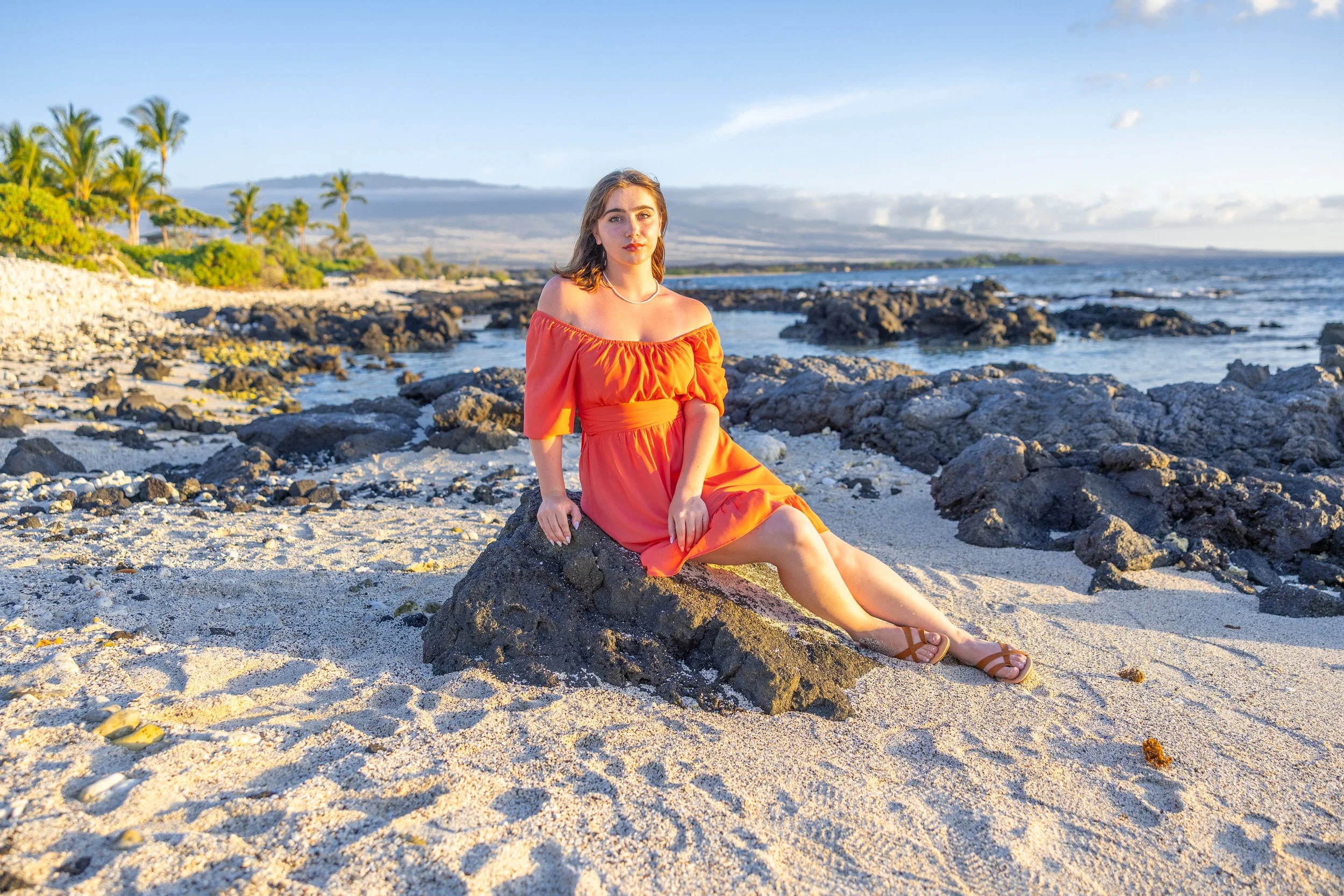 Woman in an orange dress sitting on a rock on a sandy beach, with palm trees, rocks, ocean, and sky in the background.