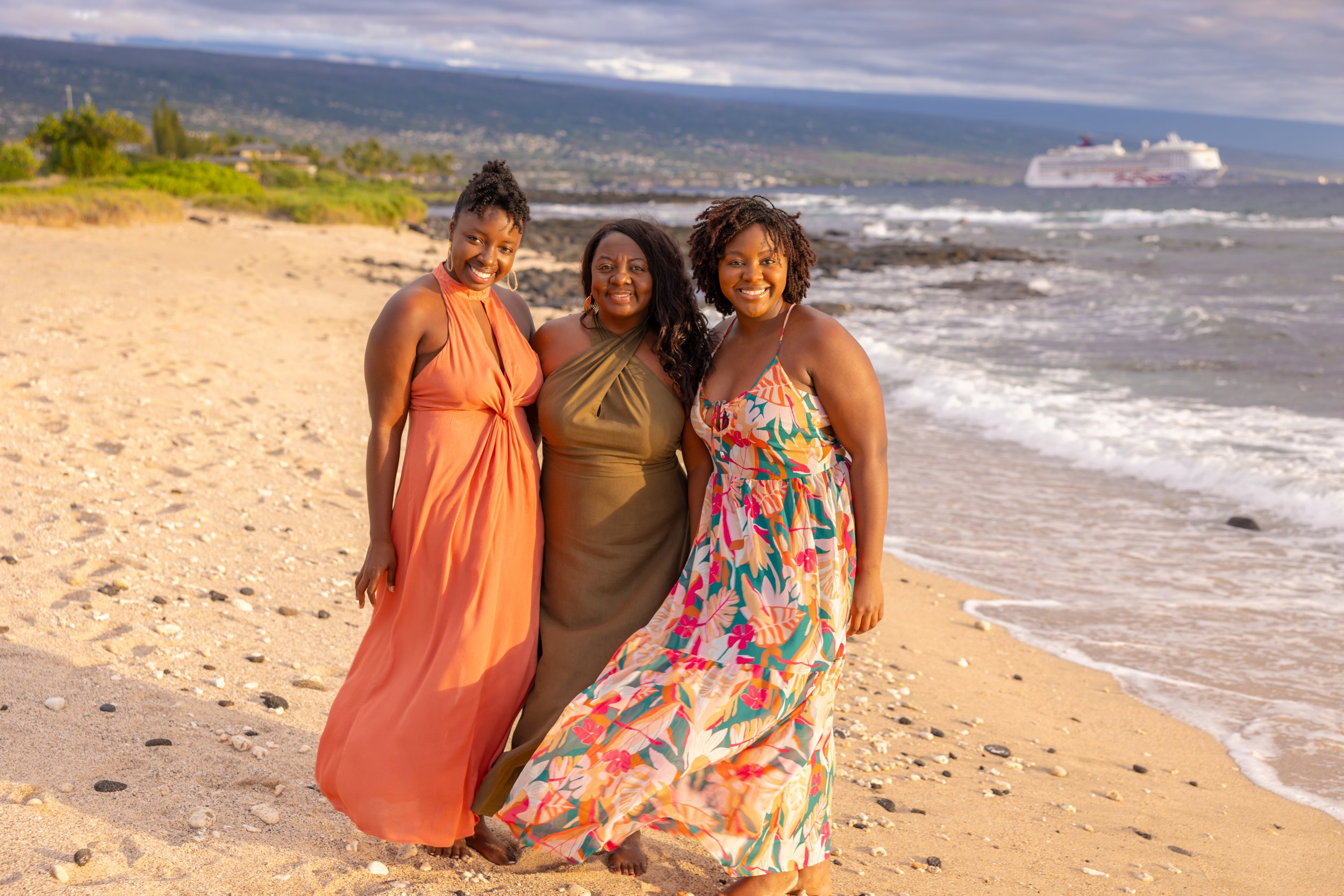 Three women smiling and posing on a sandy beach with ocean waves, a cruise ship in the distance, and mountains in the background.