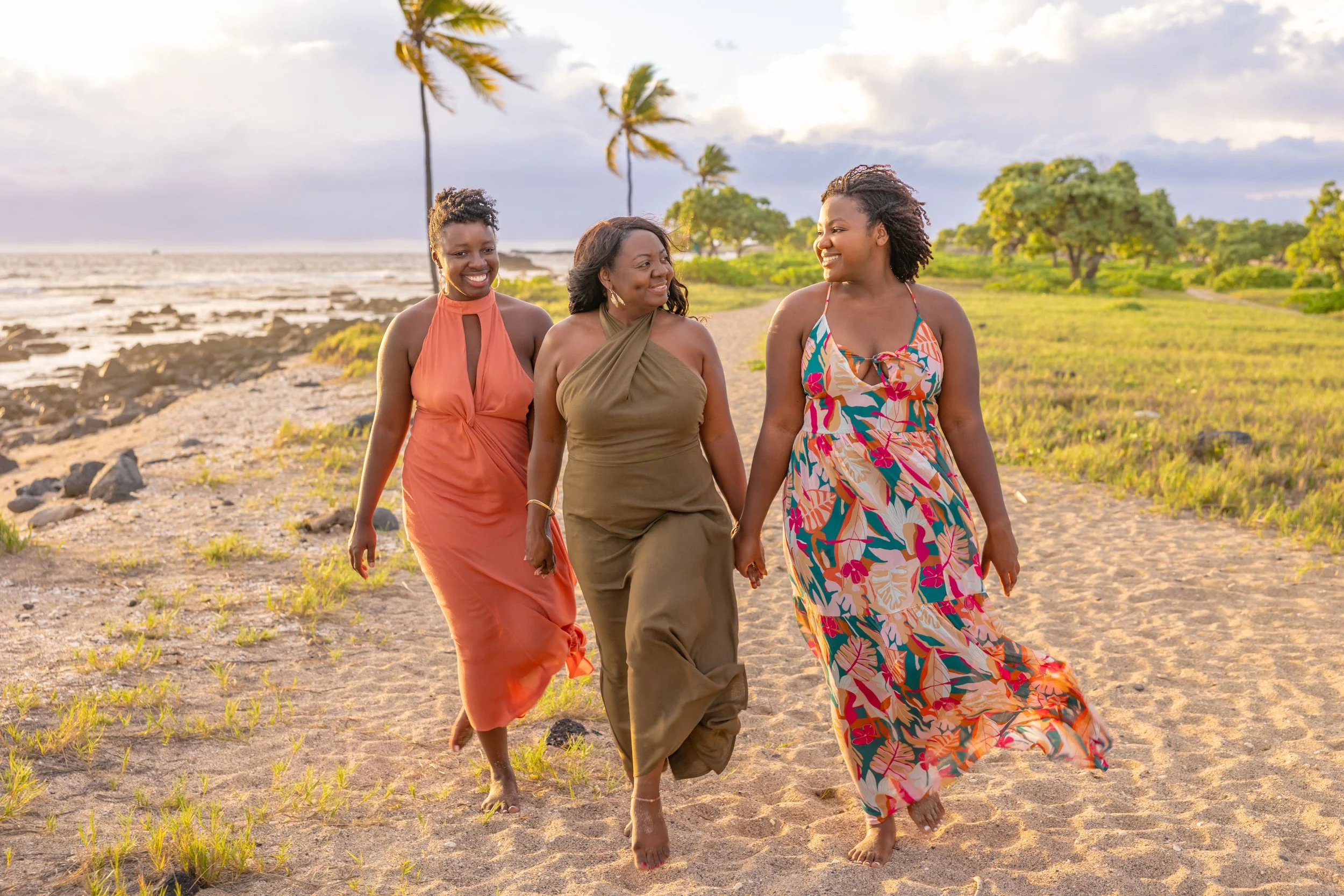 Three women walking hand in hand on a sandy beach with palm trees, green grass, and a cloudy sky in the background during sunset.