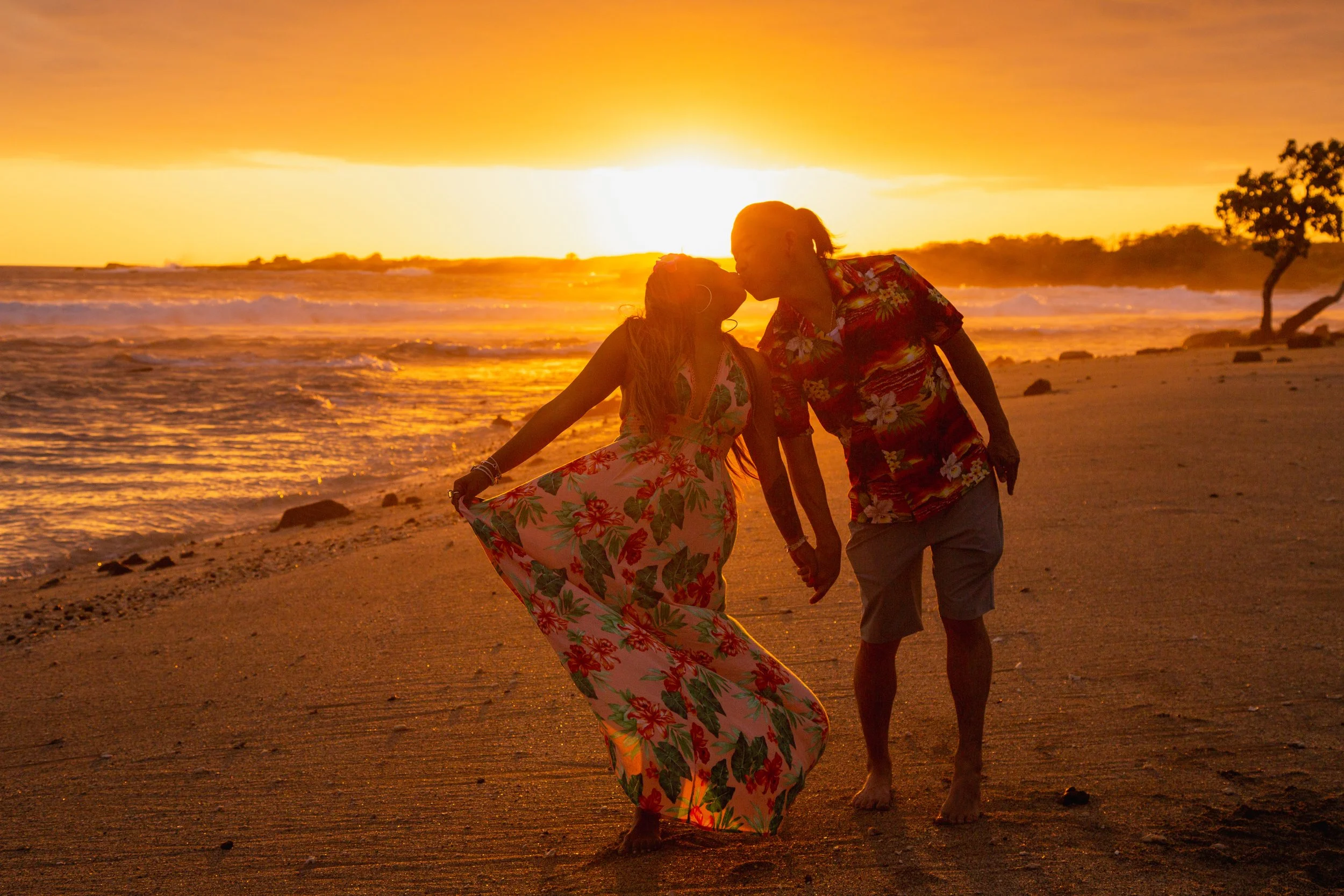 A couple holding hands and kissing on the beach at sunset, with the ocean and trees in the background.