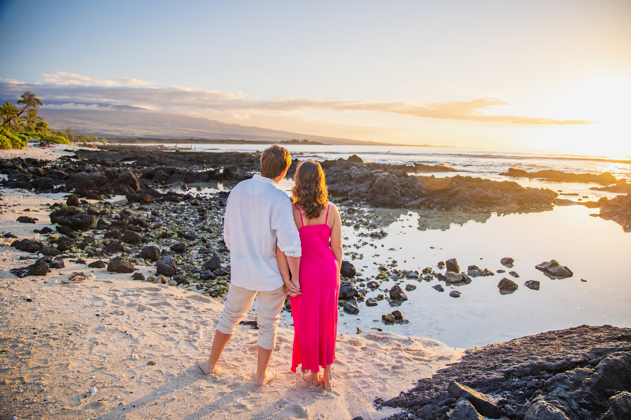 A couple holding hands standing on a sandy beach at sunset, facing the ocean with rocks and water in the background.