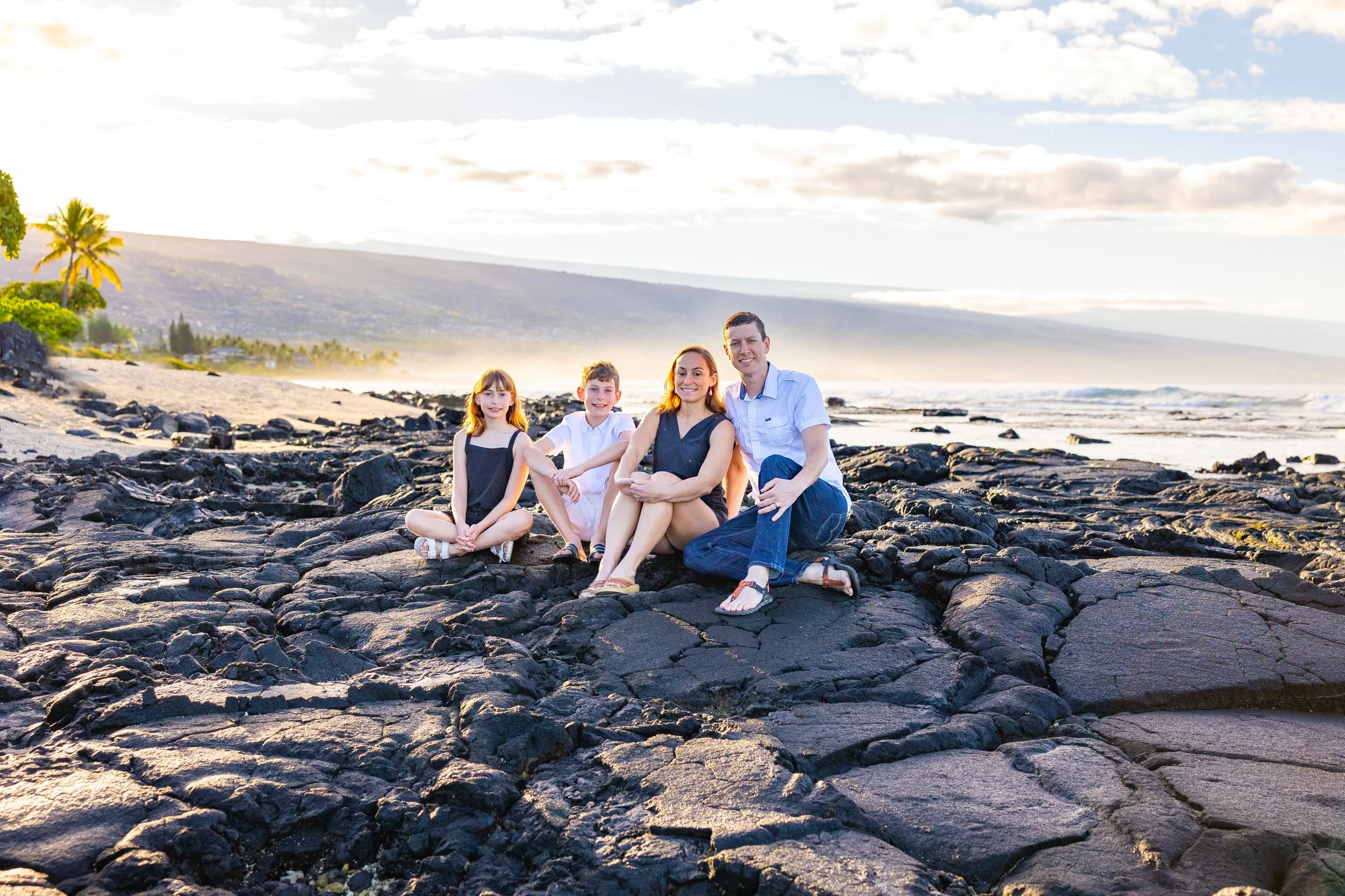 A family of four sitting on the rocky beach in the late afternoon, with the ocean, distant mountains, and partly cloudy sky in the background.