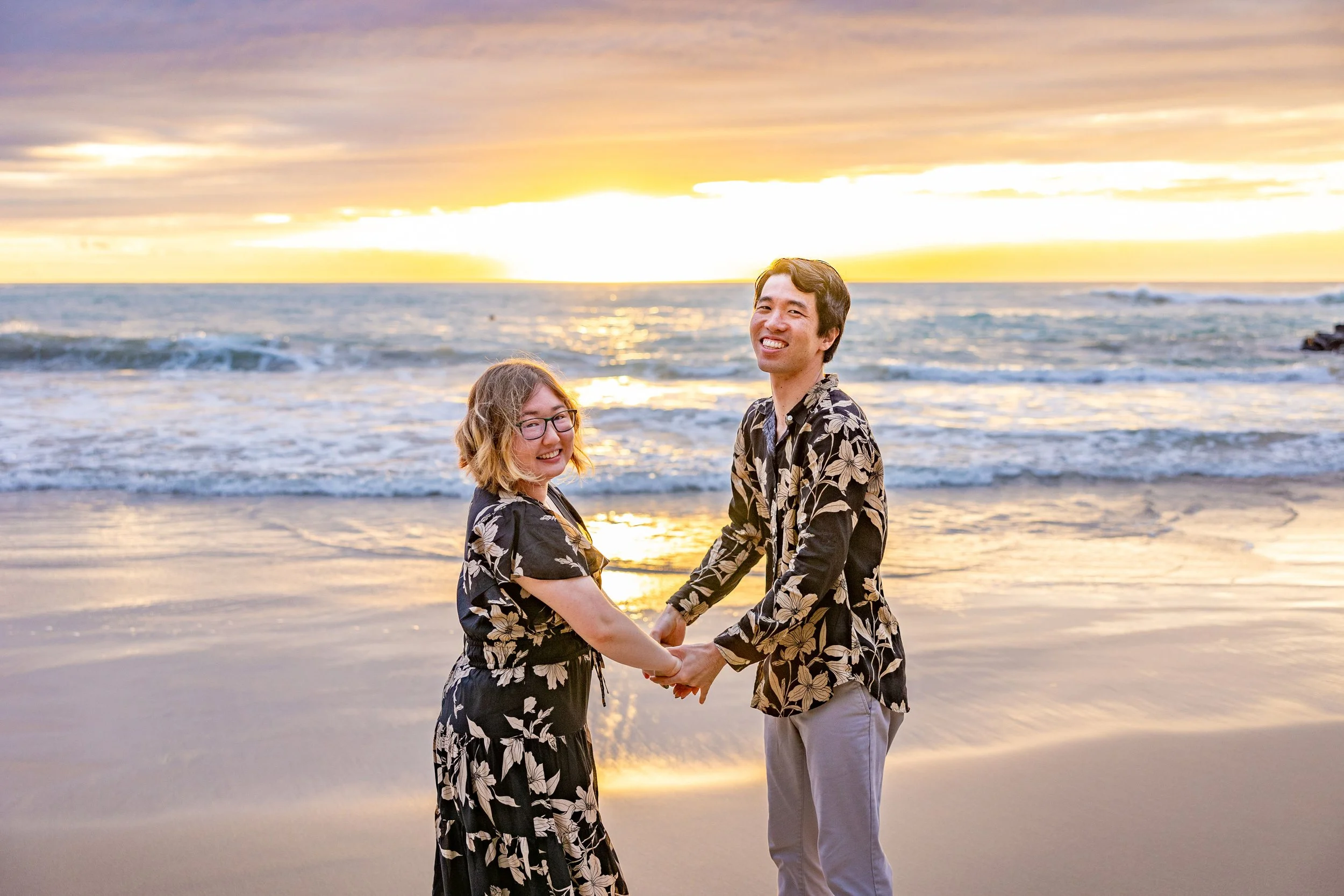 A smiling couple holding hands on the beach during a sunset, with the ocean and orange sky in the background.