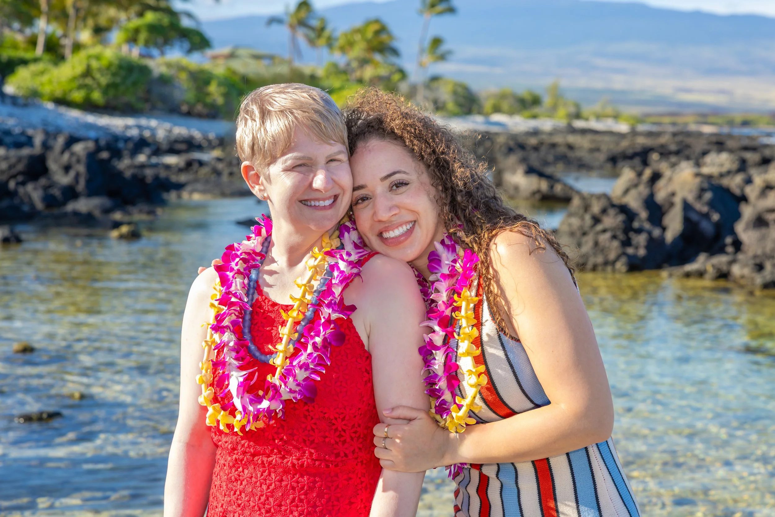 Two women smiling and hugging on a beach, wearing colorful leis.