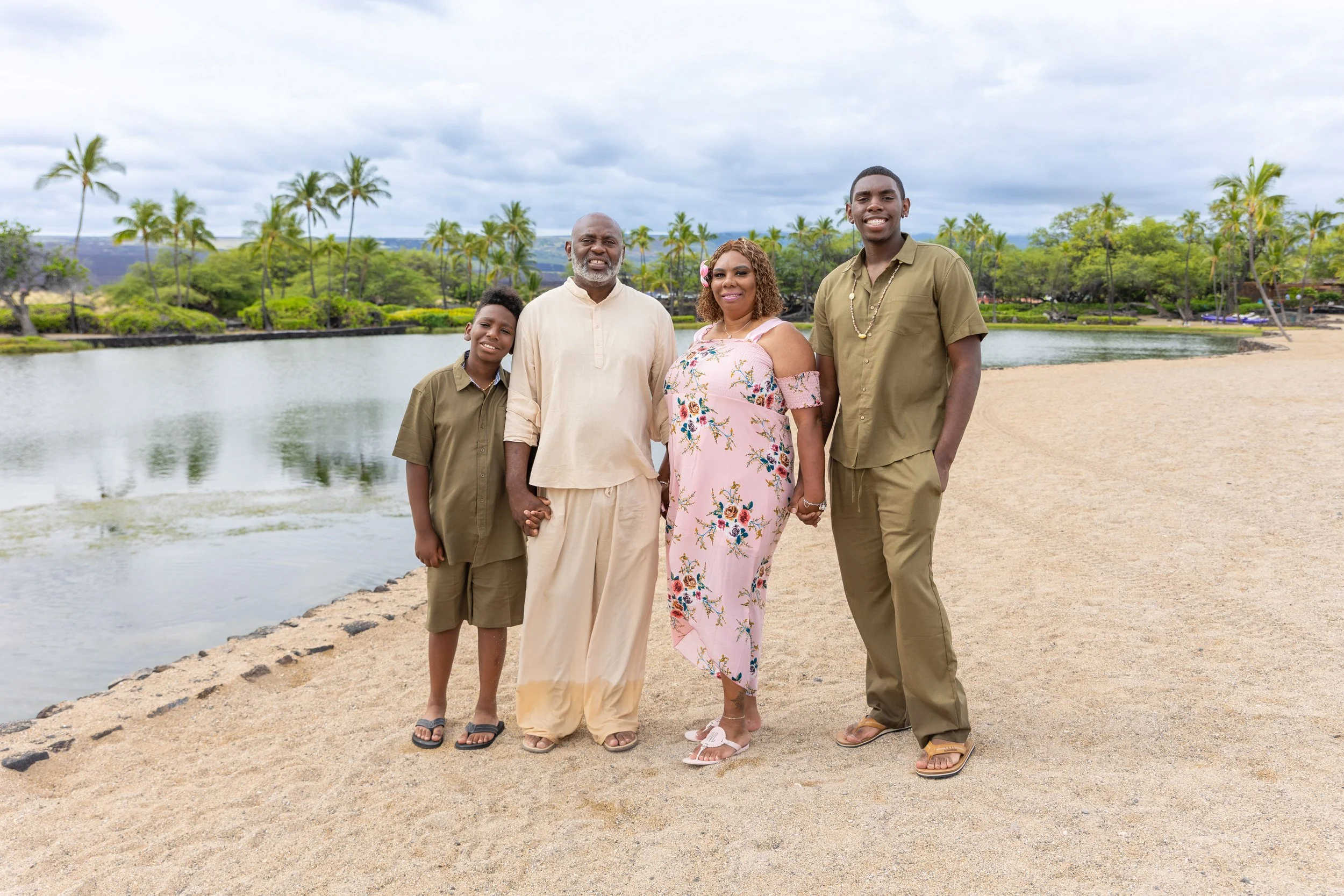 A family of five standing on a sandy beach near a body of water, with palm trees and greenery in the background on a cloudy day.