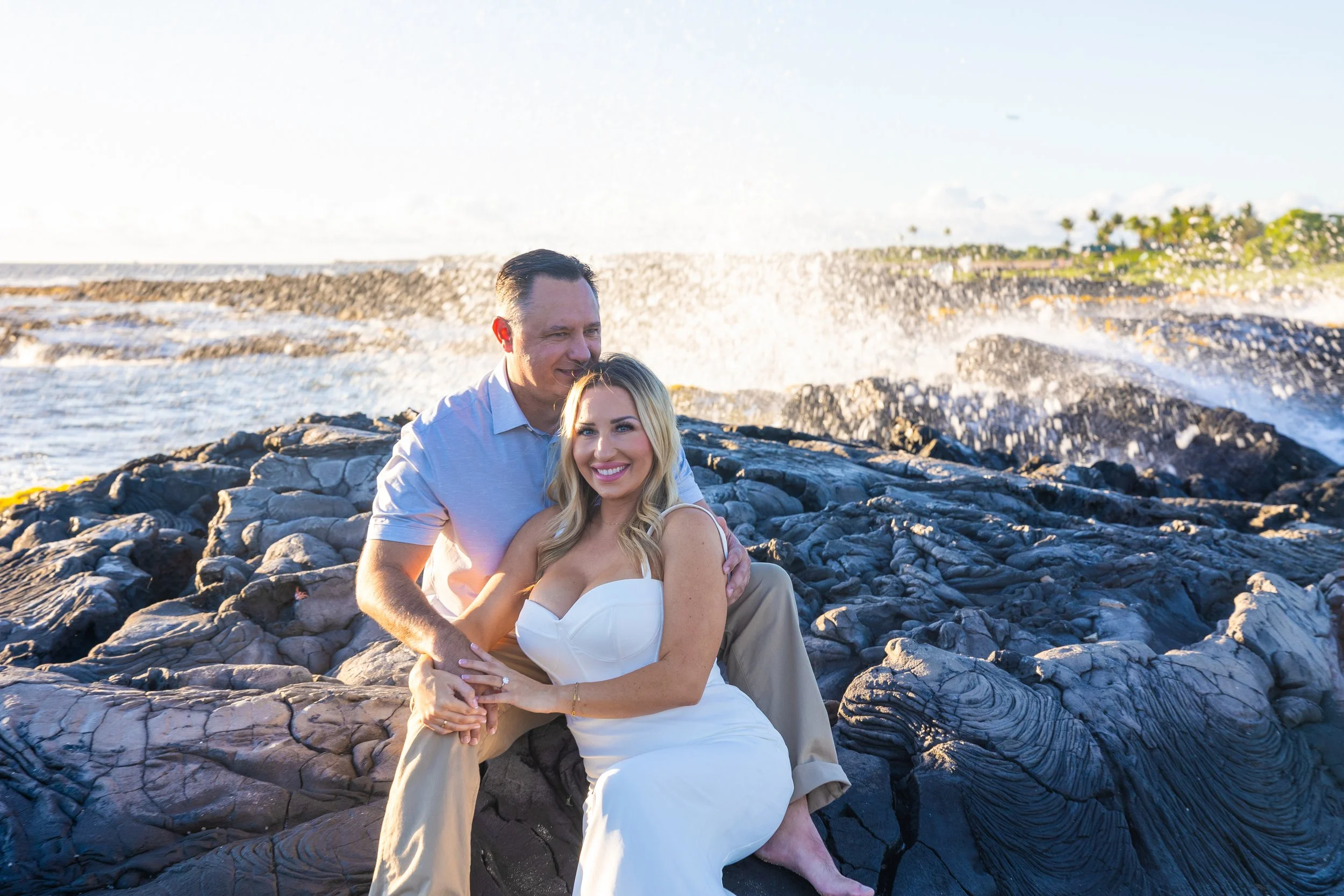 A couple sitting close together on a rocky shoreline, smiling, with waves crashing behind them and a hint of trees in the distance.