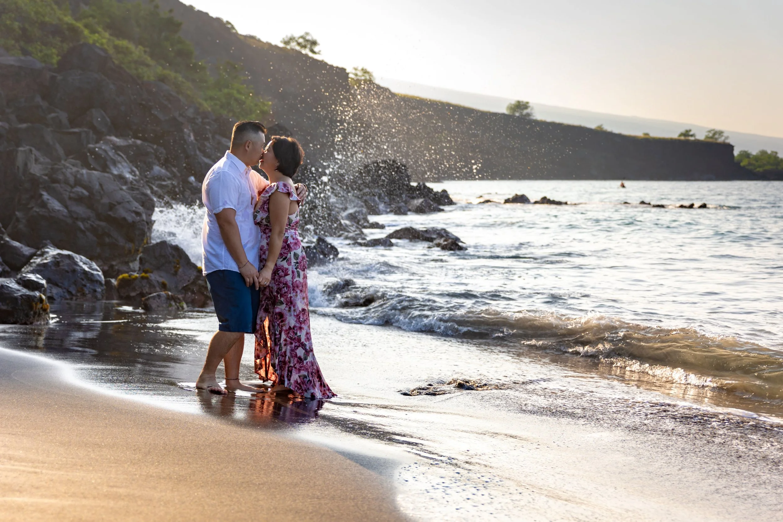 A couple stands close together on a beach, holding hands and kissing near the shoreline during sunset, with waves and rocks in the background.