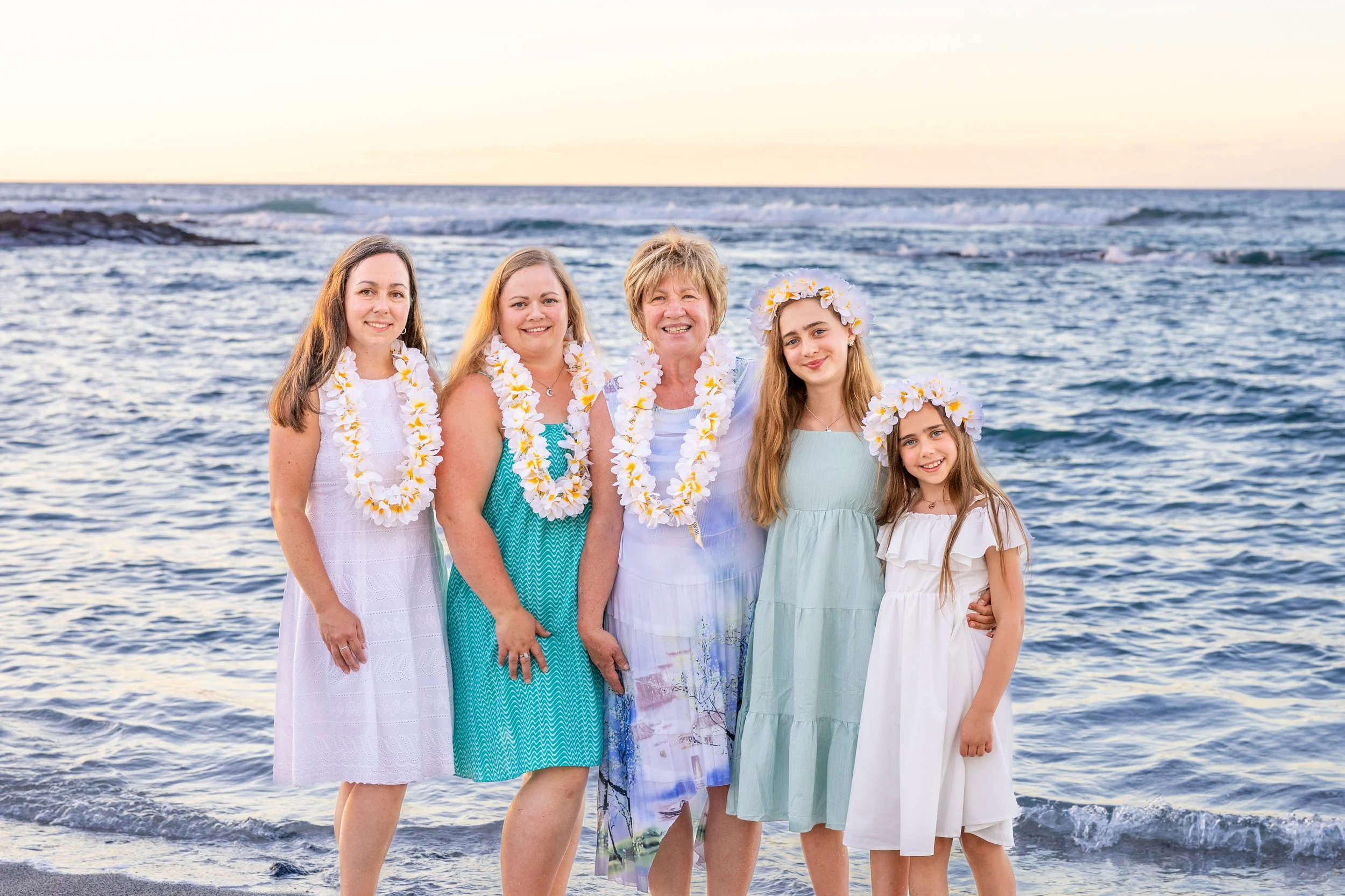 Six women and girls in pastel dresses and flower leis standing on a beach at sunset, smiling at the camera.
