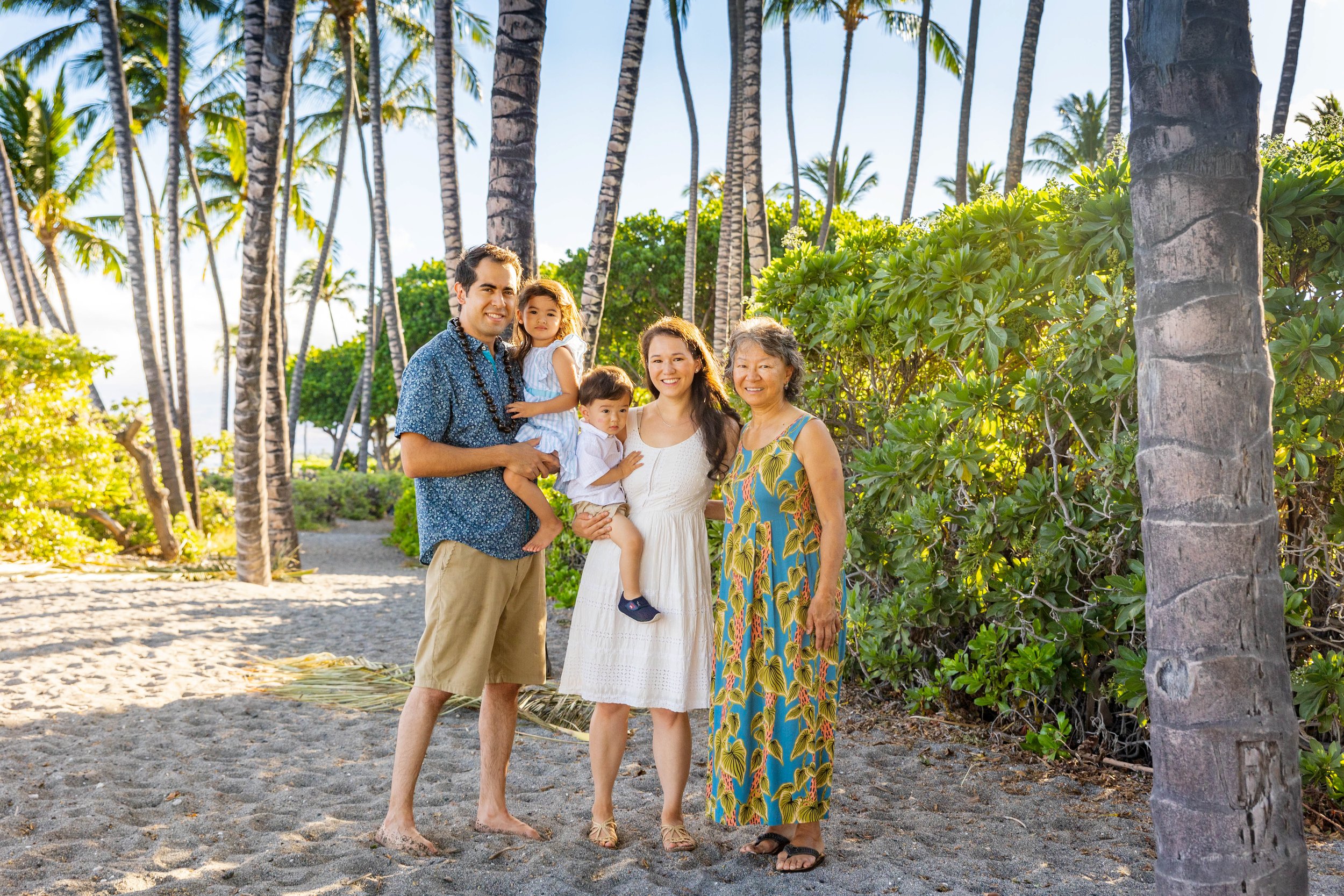 A family of five standing on a sandy beach among palm trees, smiling and enjoying a sunny day, with lush green foliage around.