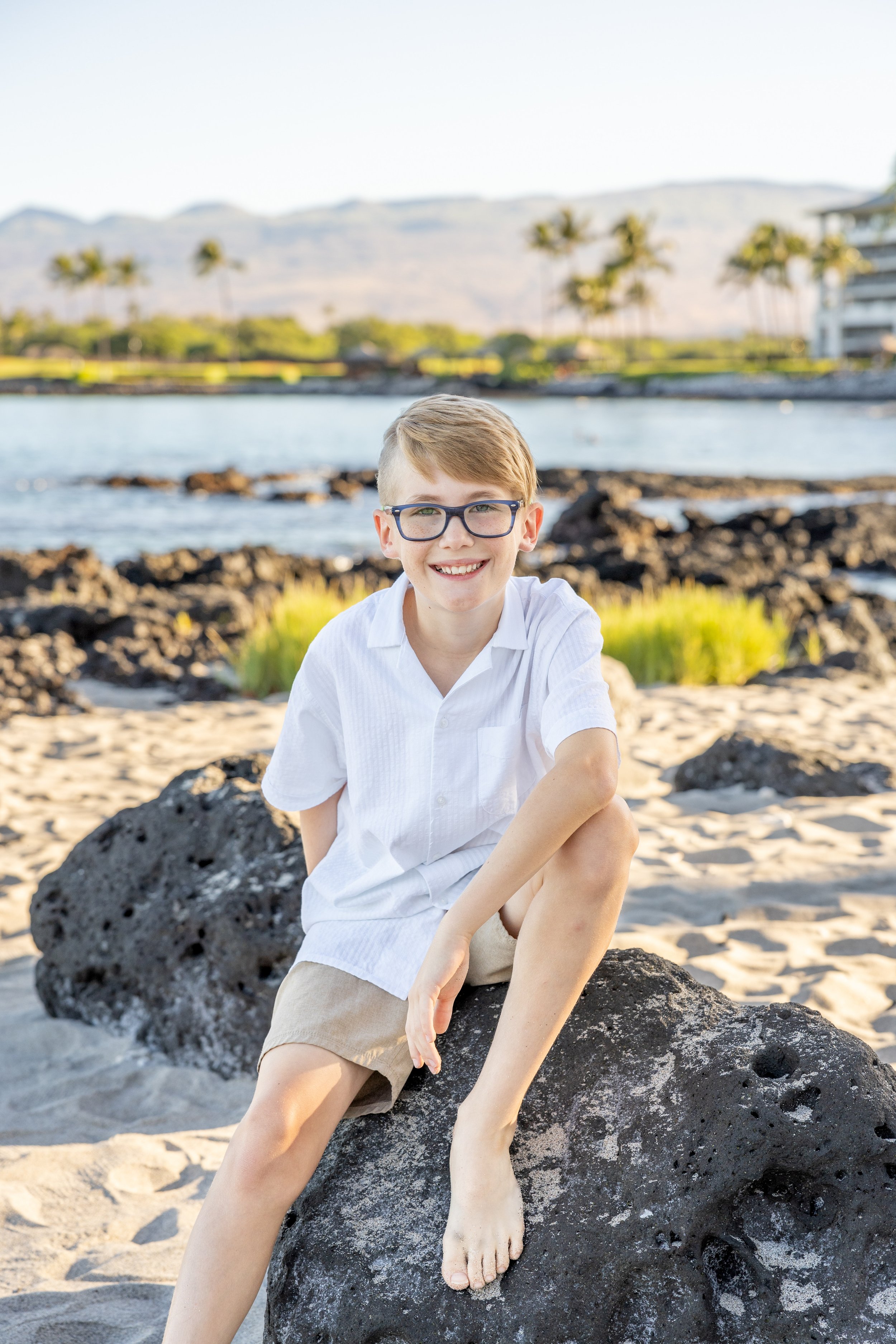 A young boy with glasses, wearing a white shirt and beige shorts, sitting on a large black volcanic rock on a sandy beach with water, rocks, green plants, and palm trees in the background.