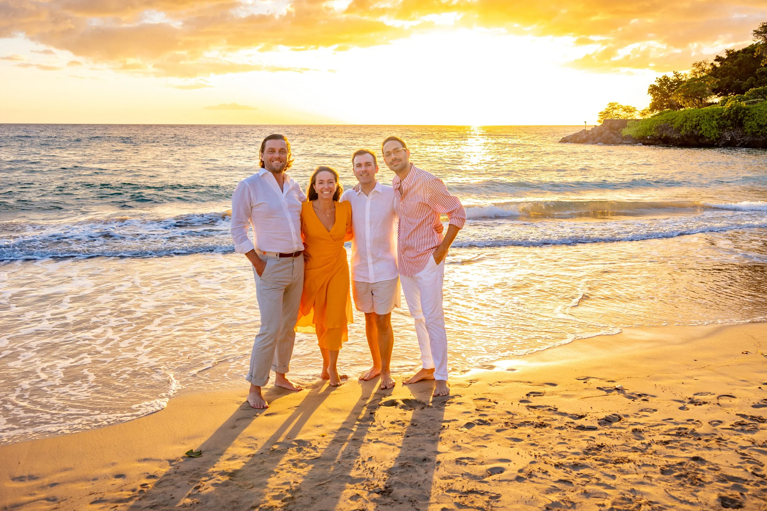 Four friends standing on a beach at sunset, smiling and posing for a photo.