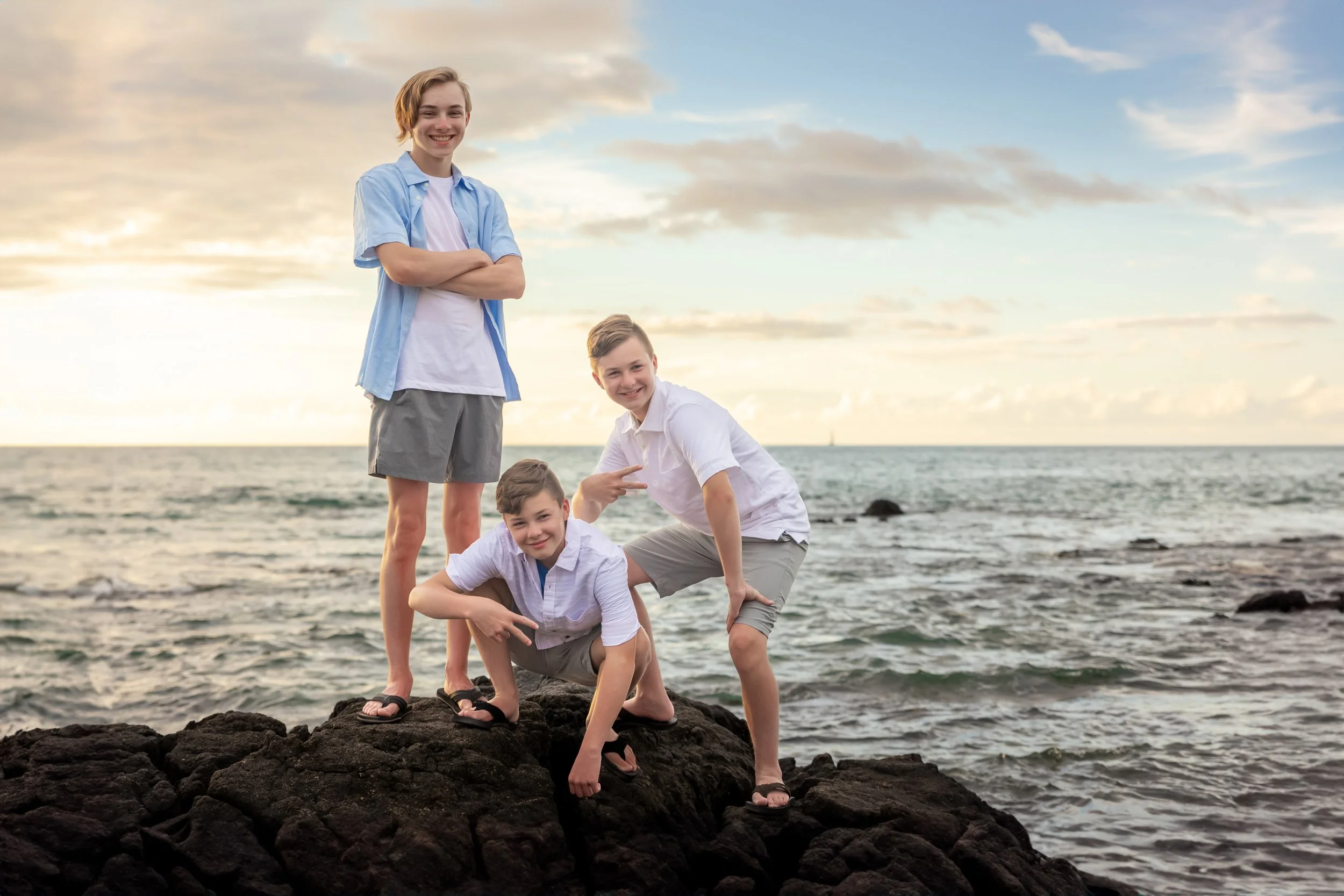 Four kids wearing casual clothes on rocks near the ocean at sunset, smiling and posing.