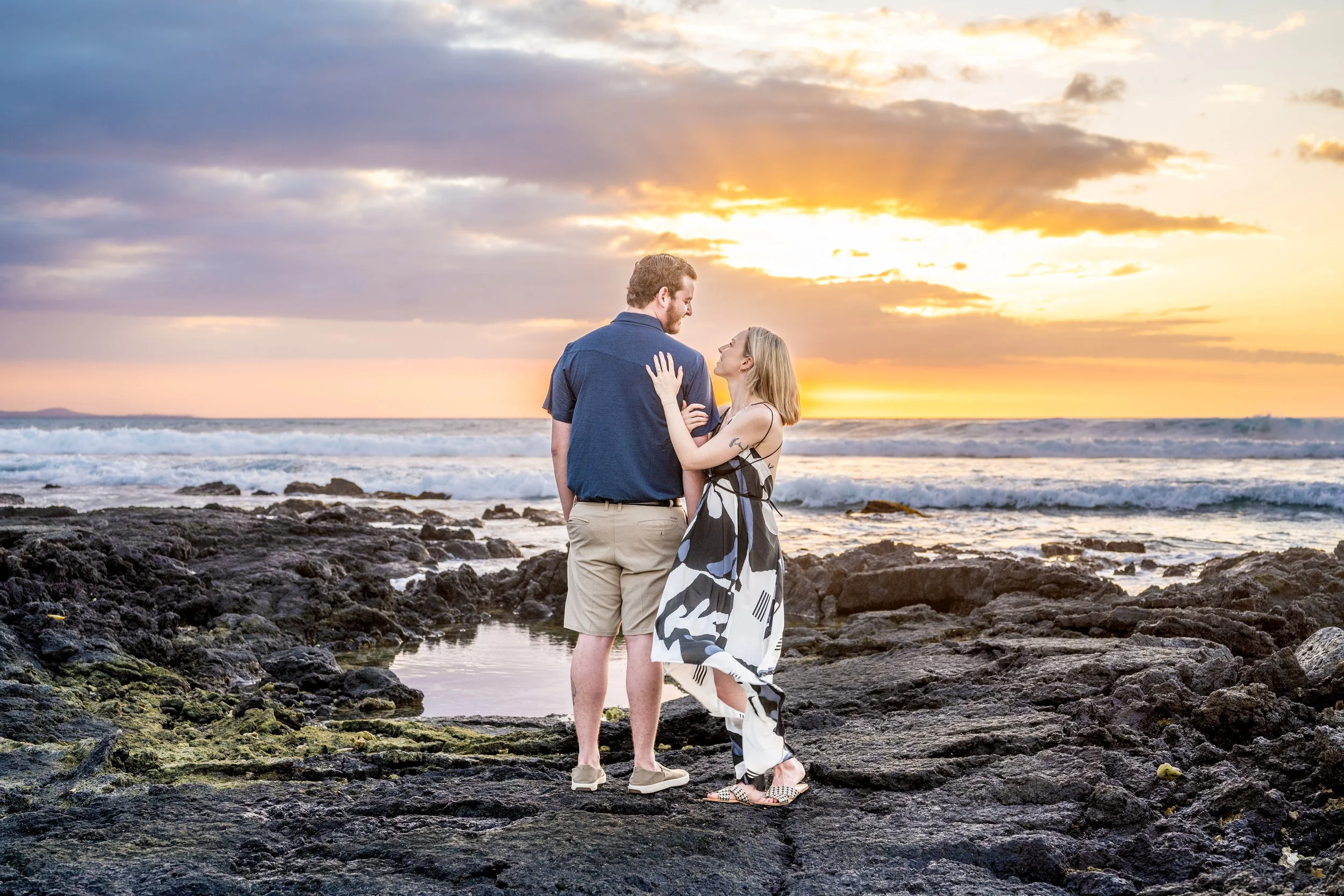 A couple stands on a rocky beach at sunset, gazing at each other, with waves in the background.