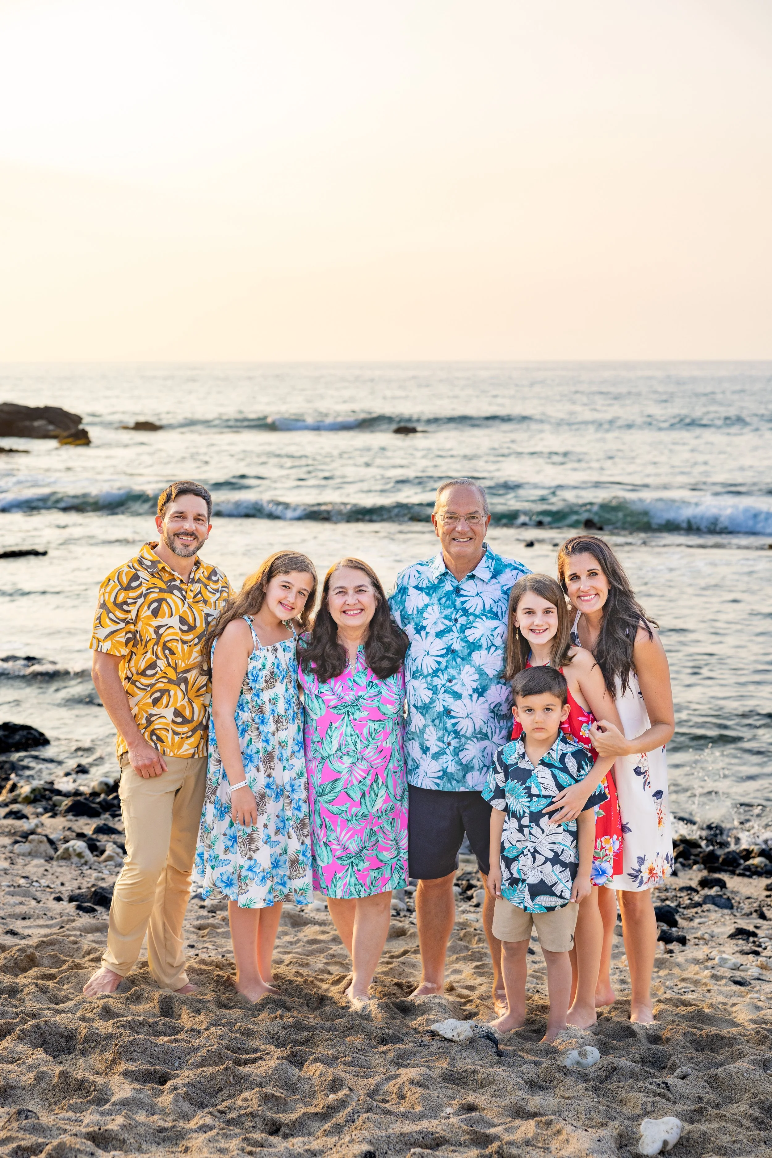 Family standing on a sandy beach with ocean in the background, all dressed in colorful, tropical patterned summer clothes, smiling at the camera.