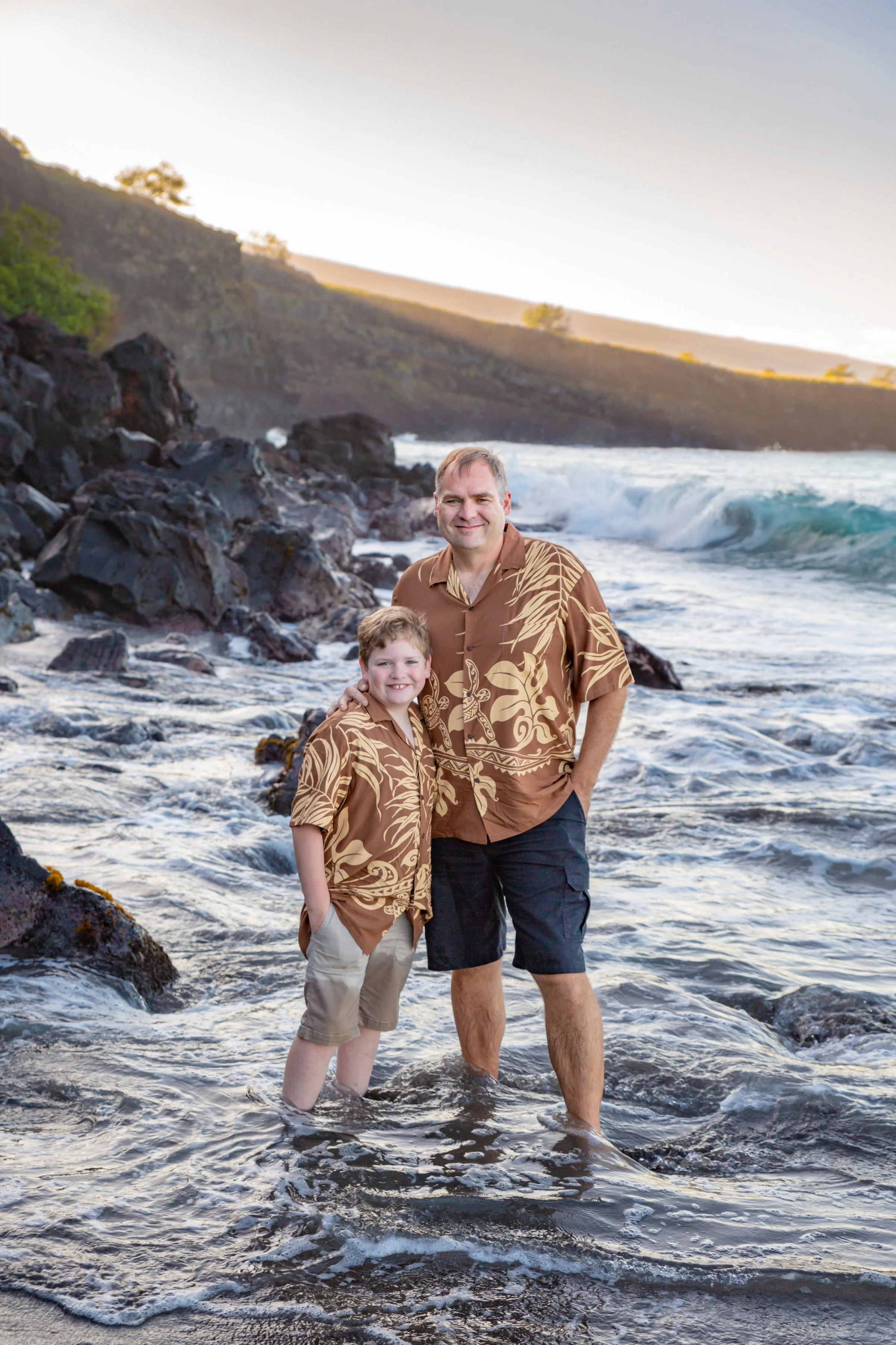 A father and son standing in the ocean at sunset, both wearing matching Hawaiian shirts, smiling at the camera.