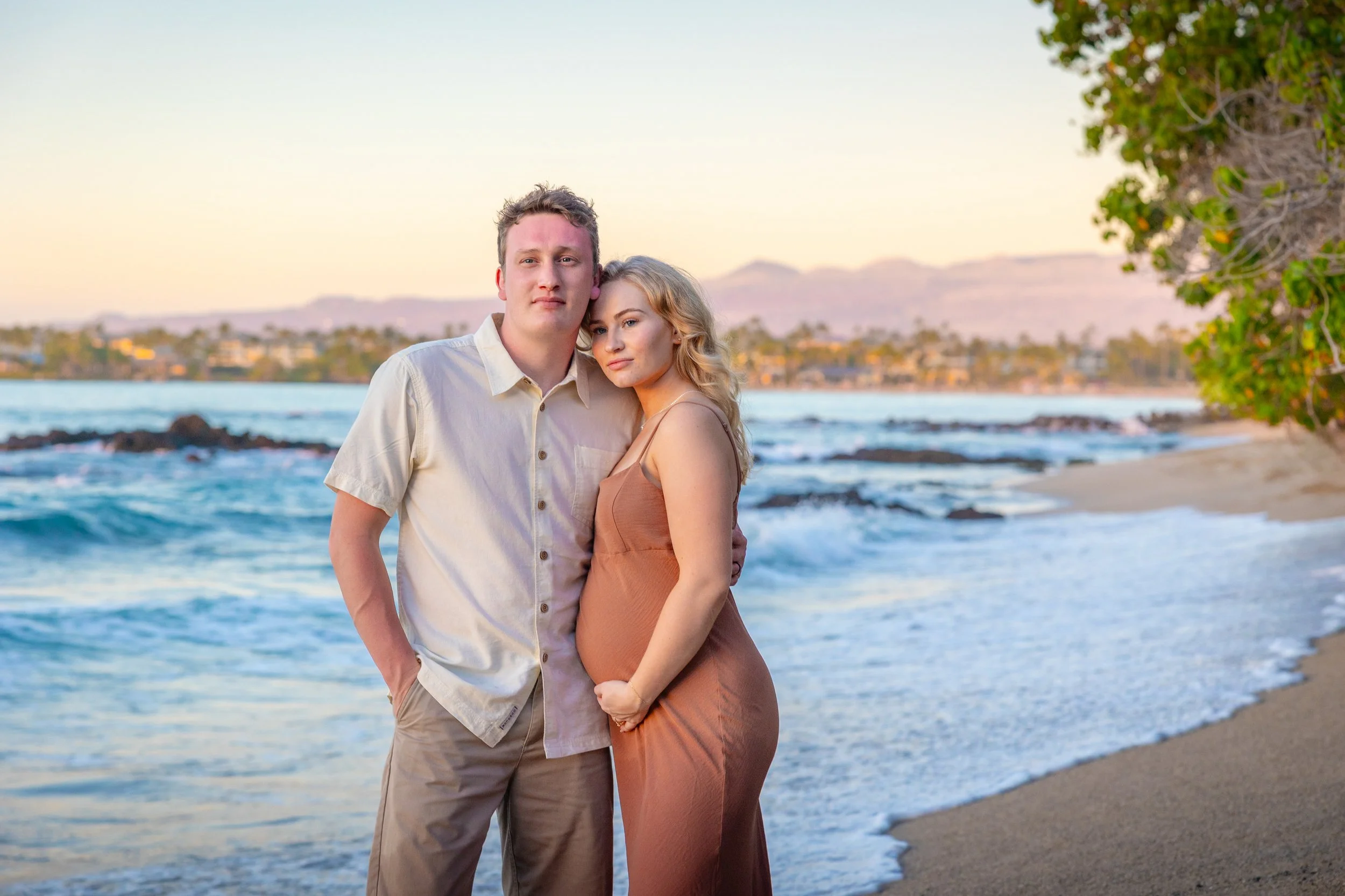 A pregnant woman and a man standing on a beach during sunset, with waves and trees in the background.