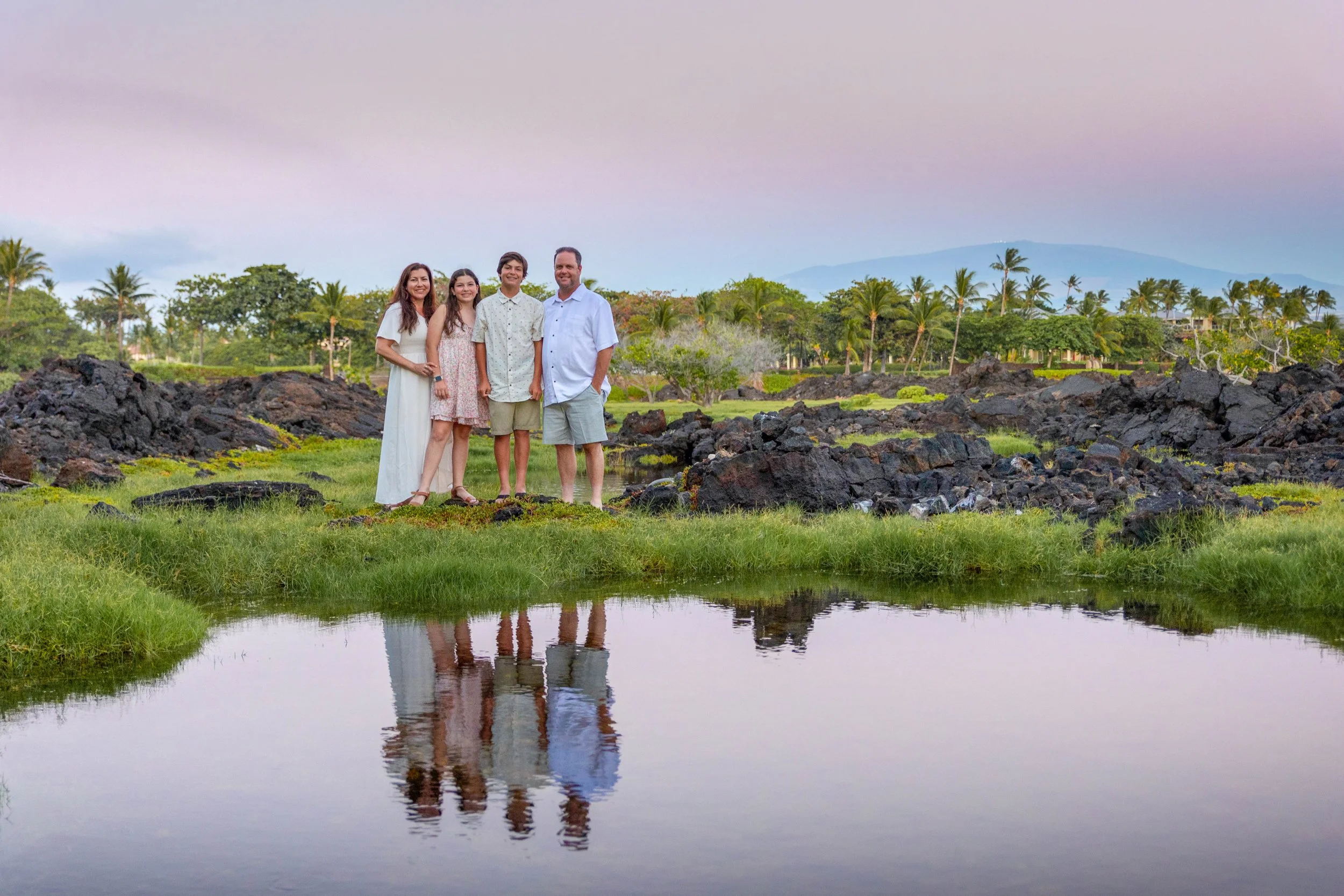 A family of four standing near a pond with reflections, lush greenery, palm trees, and mountains in the background during sunset.