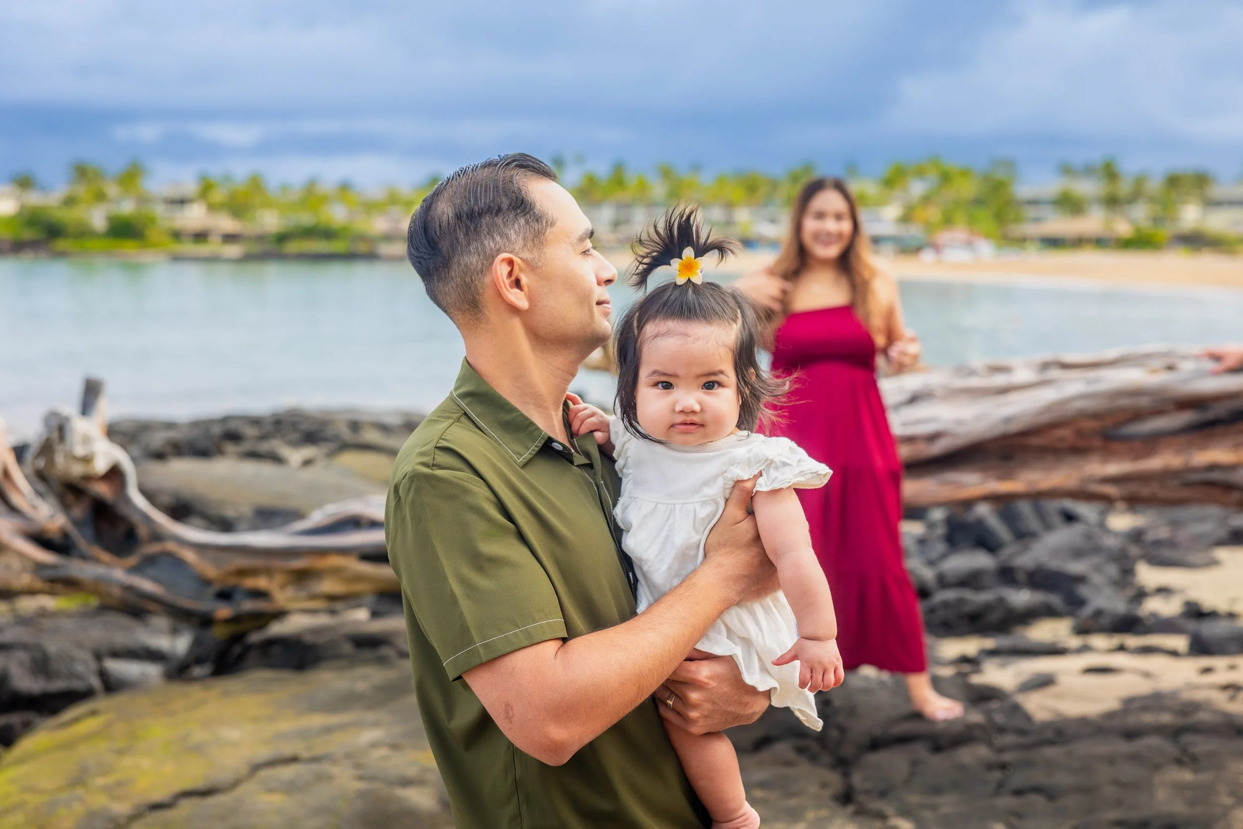 Family at the beach with a man holding a young girl with a flower in her hair, and a woman in a red dress standing in the background near logs and rocks by the water.