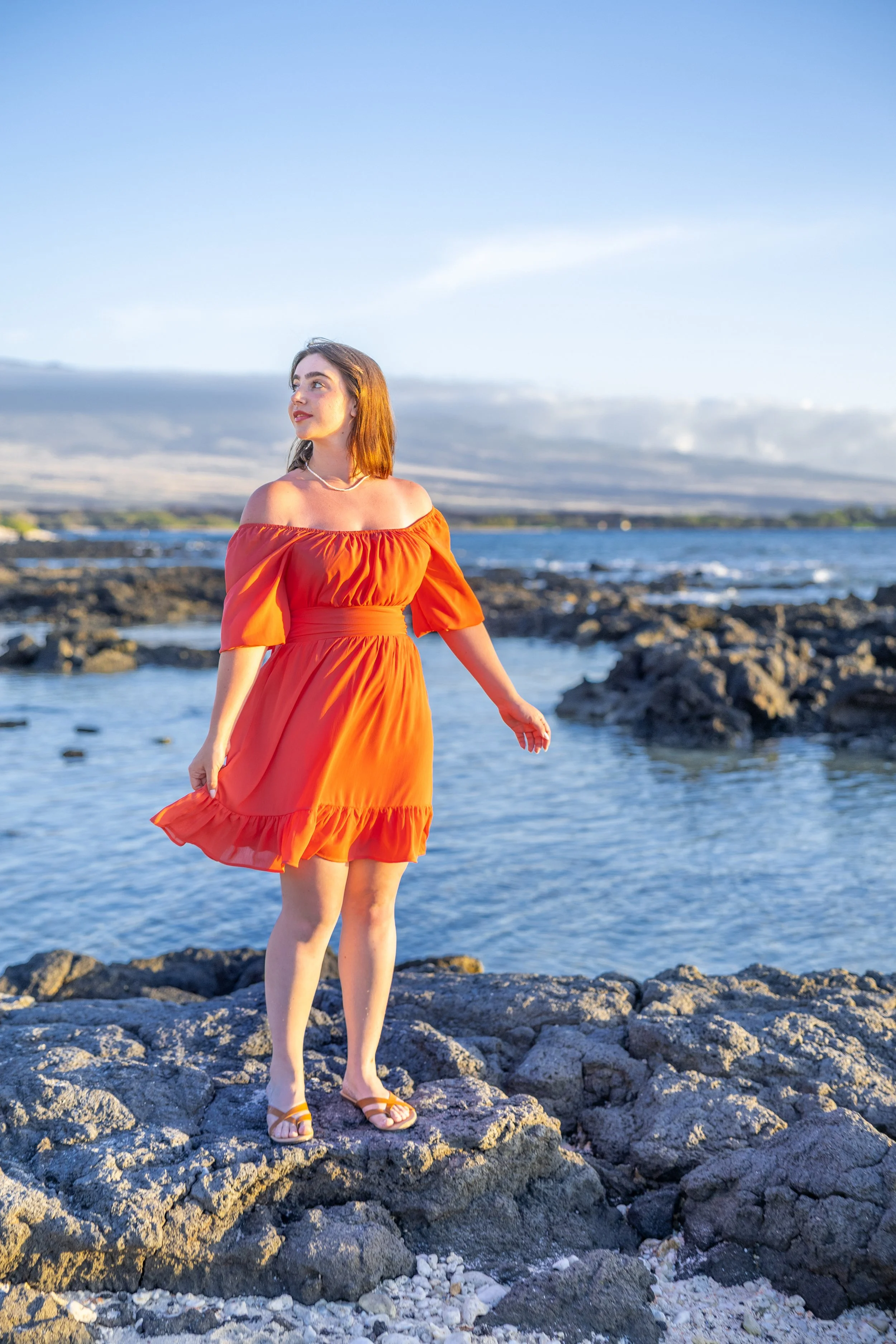 A woman in an orange dress standing on rocks by the ocean with a scenic coastal background.