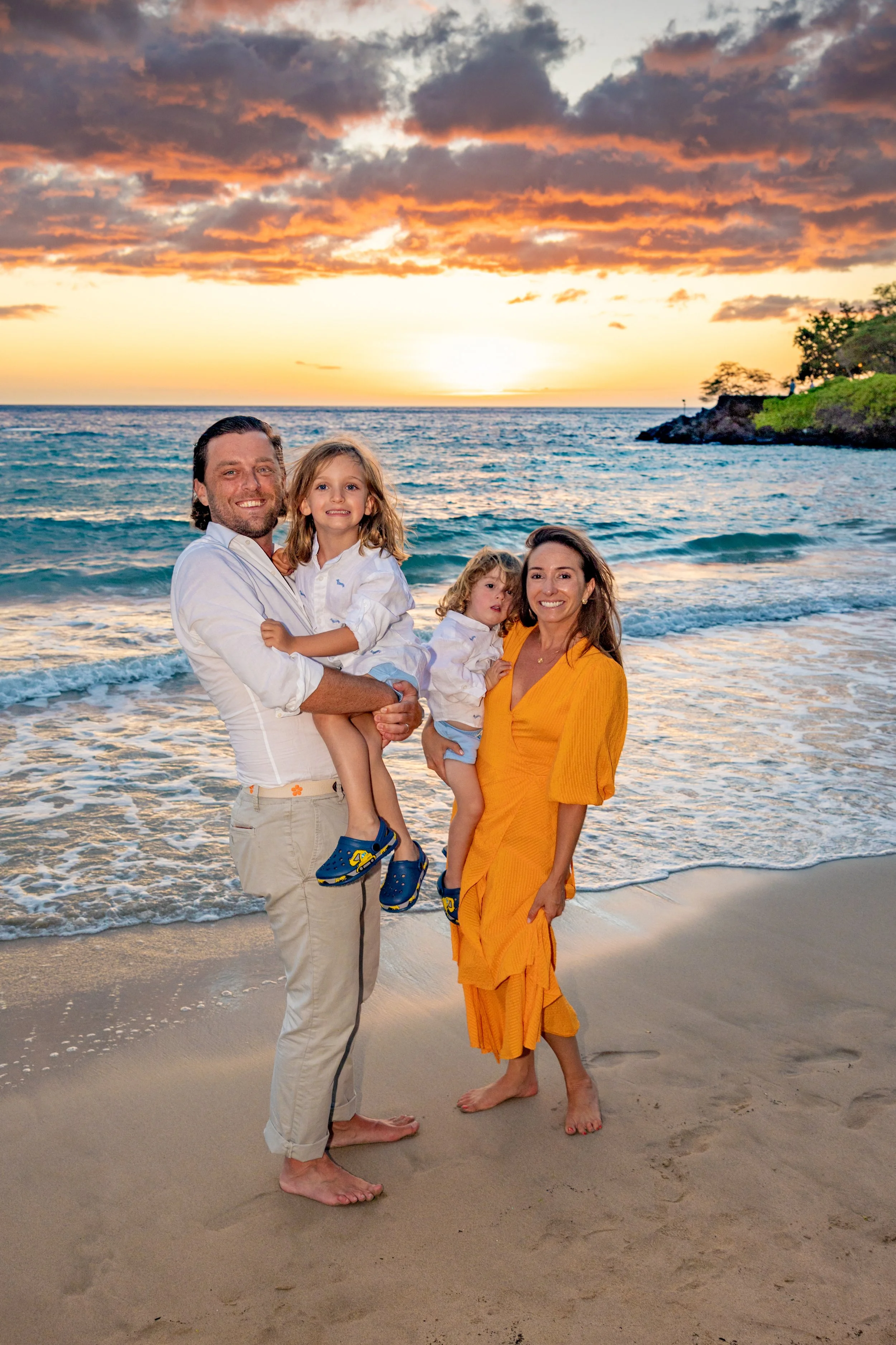 A family of four standing on a sandy beach during sunset. The father is holding one daughter, and the mother is holding the younger daughter. The ocean and a partly cloudy sky with a colorful sunset are in the background. All family members are smili