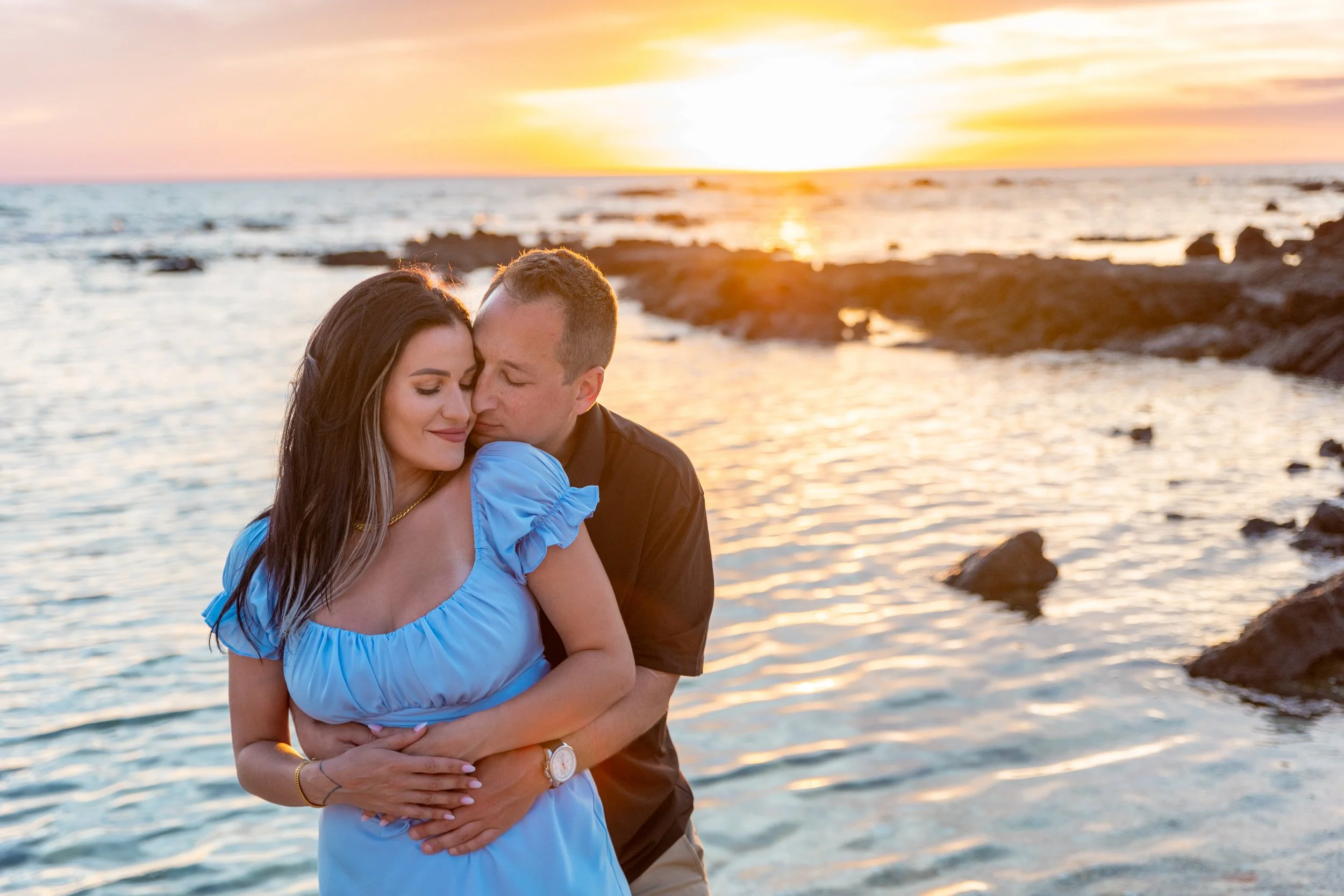 A couple embracing at sunset on a beach with rocks and calm water.