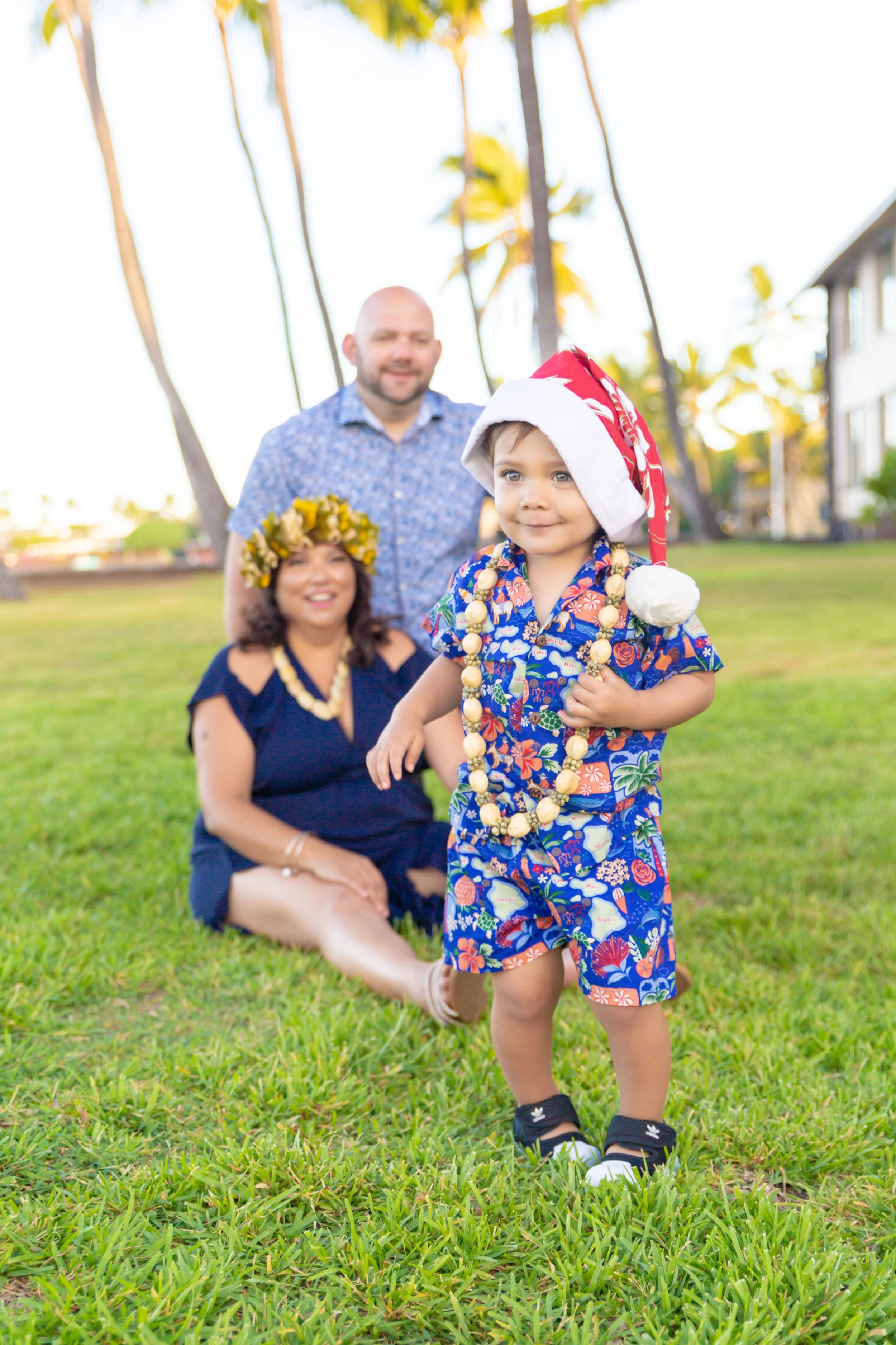 A young boy in colorful Hawaiian shirt, shorts, and sandals, wearing a Santa hat and flower necklace, walking on grass in front of a family with tropical background and palm trees.