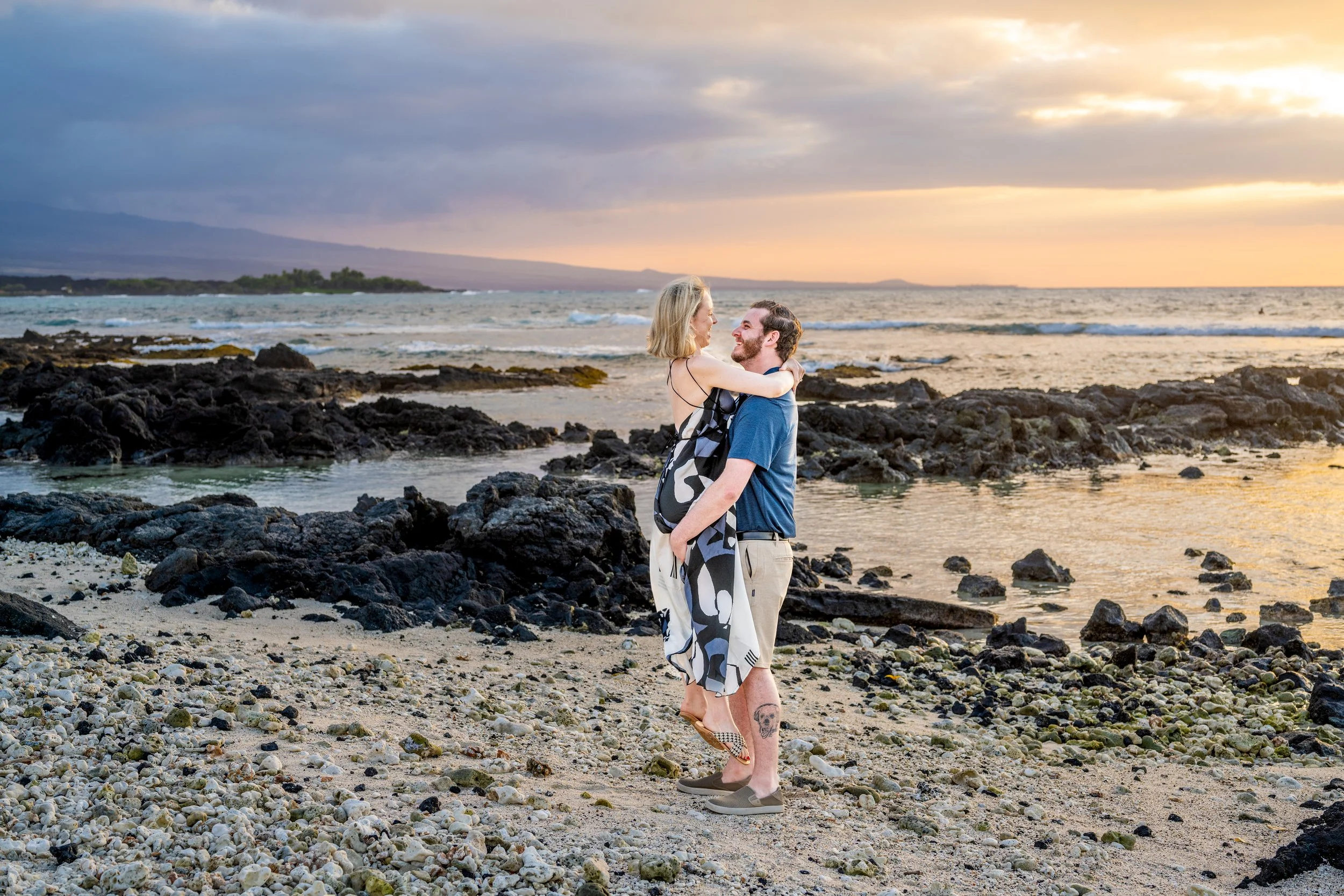 A romantic couple embracing on a rocky beach at sunset, with the woman lifted in the man's arms, overlooking the ocean.