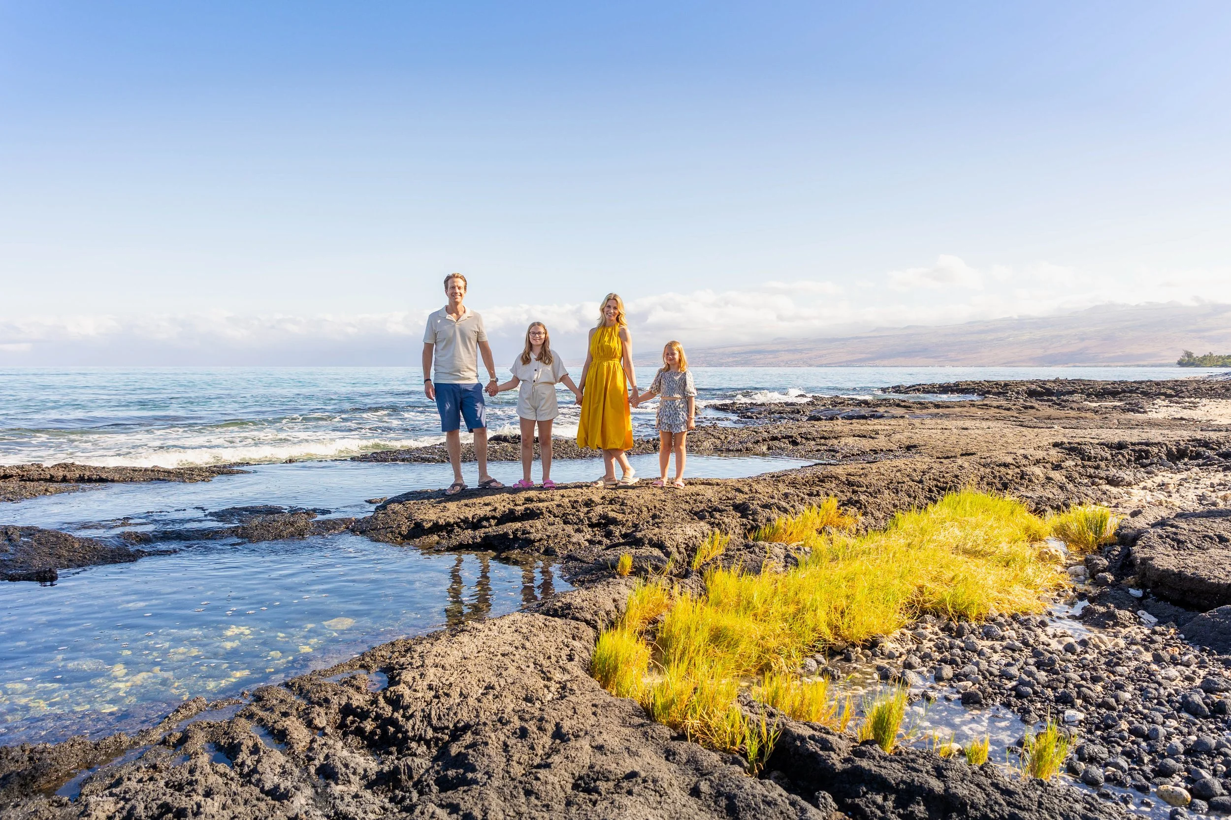 Family of four standing on a rocky shoreline by the ocean during daytime, holding hands and smiling, with mountains in the background.