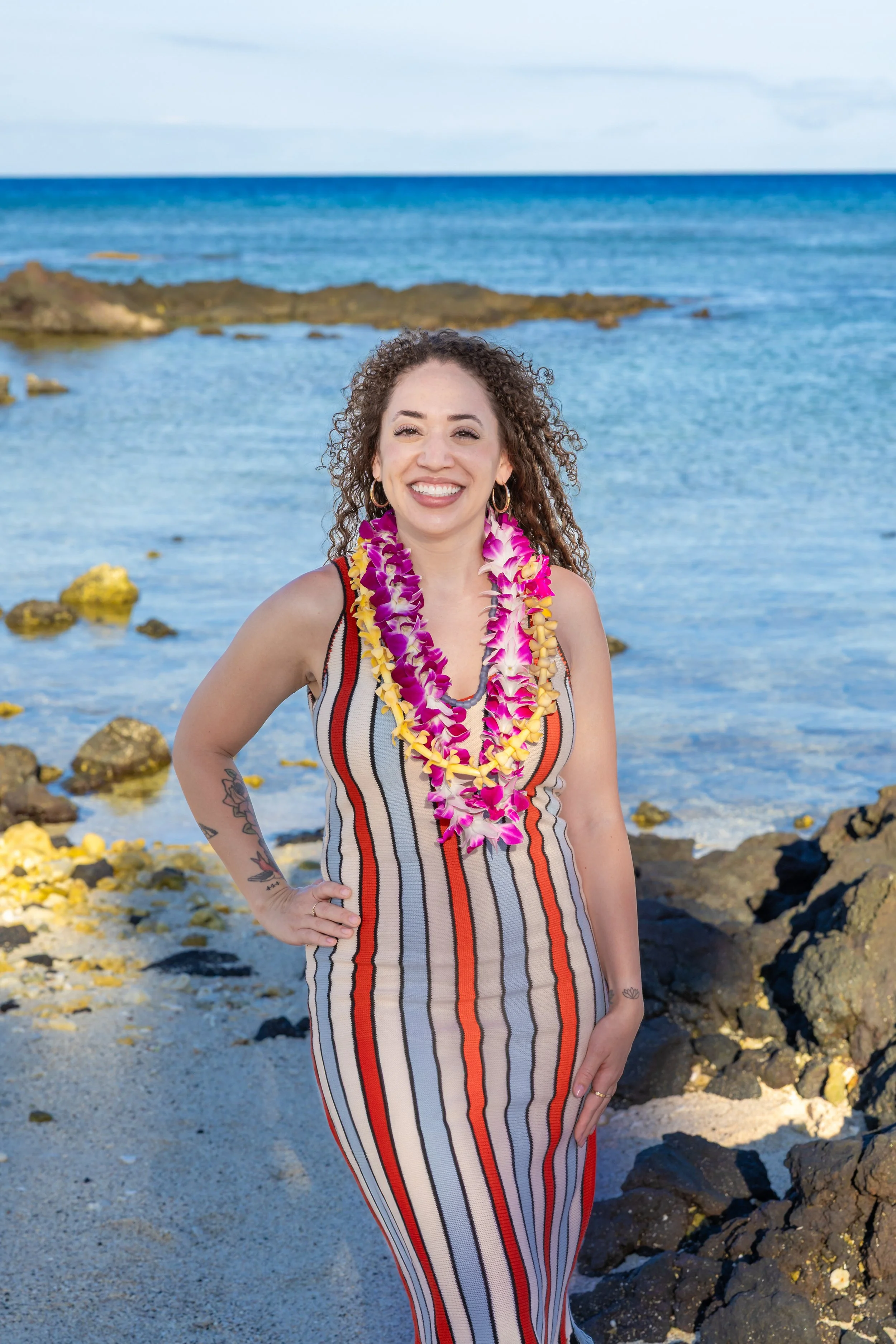 A woman standing on a rocky beach with the ocean in the background, wearing a striped sleeveless dress and colorful leis, smiling for the camera.