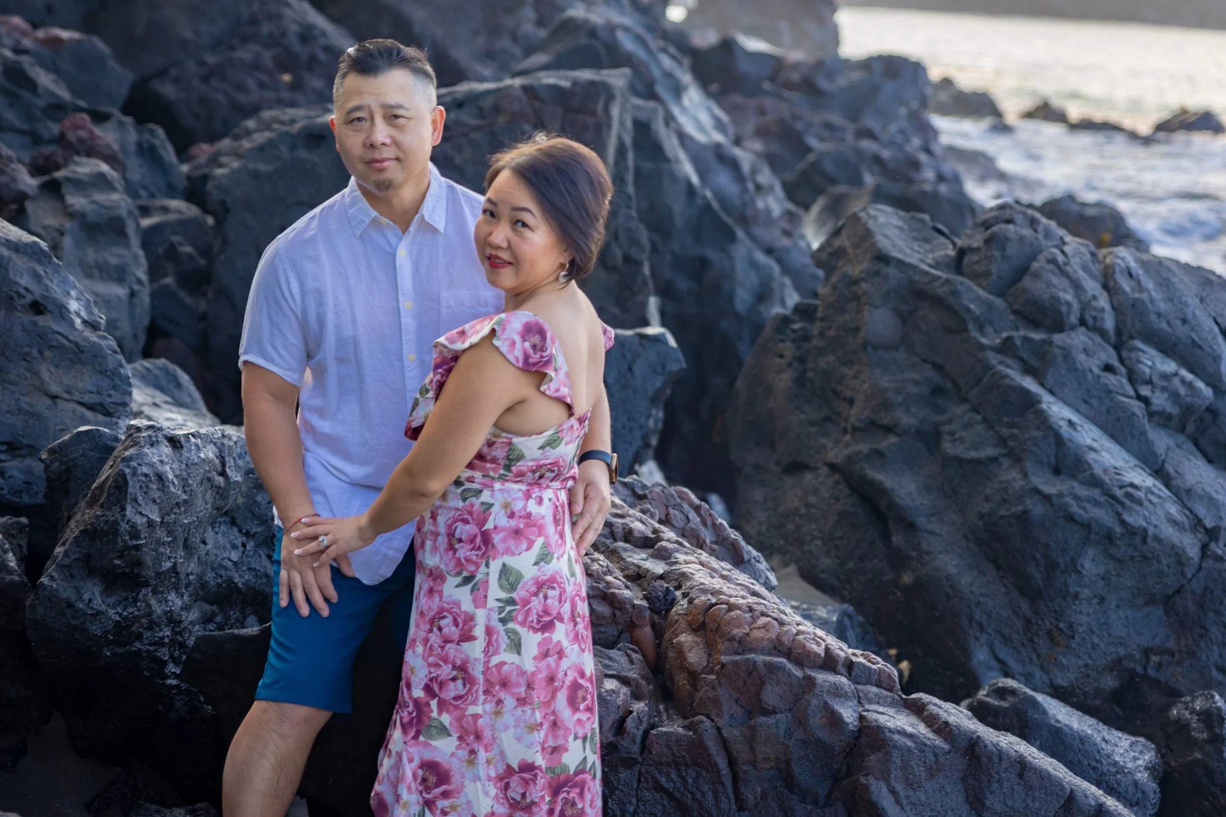 A couple standing on rocks by the ocean, with the man wearing a white short-sleeved shirt and blue shorts, and the woman wearing a pink floral dress, both looking at the camera.