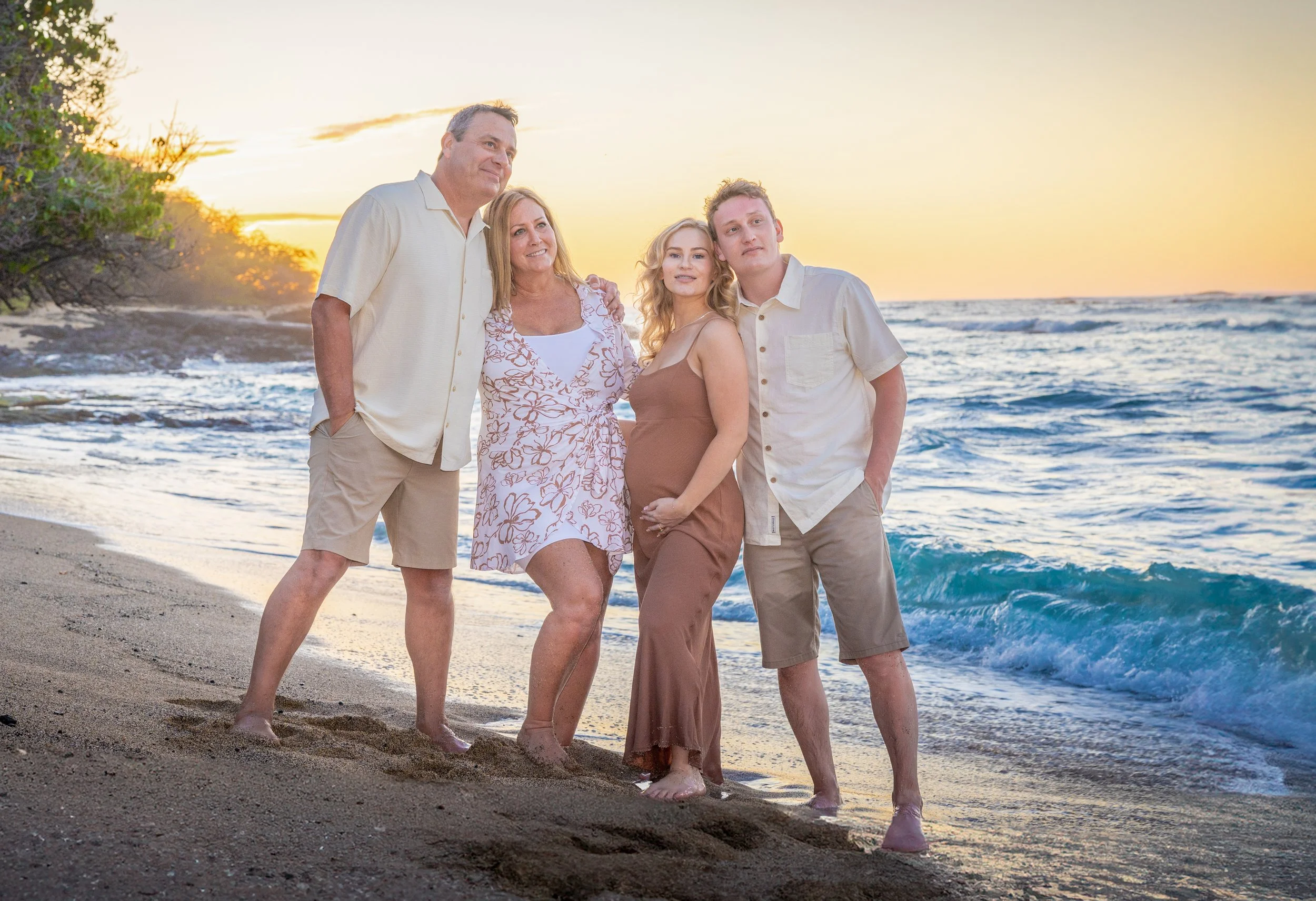 Group of five people standing on a beach at sunset, posing for a photo. They include two men, two women, and one pregnant woman. The ocean waves are visible behind them, and the sky has a warm, golden glow.